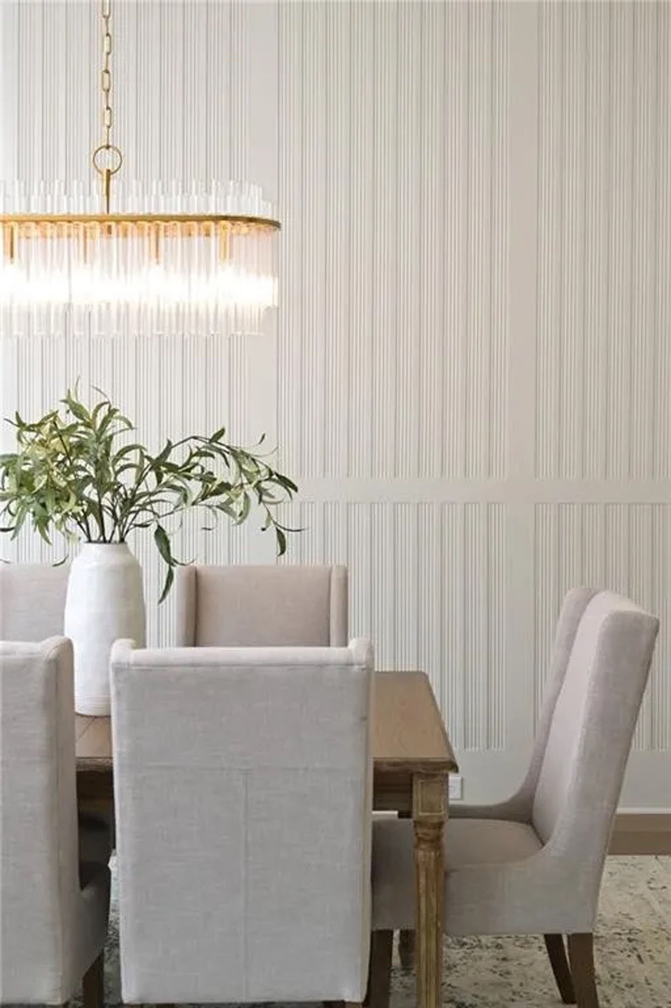 White dining table with beige upholstered chairs, a large white vase with green leafy plant, chandelier hanging above, textured light-colored wall in background.