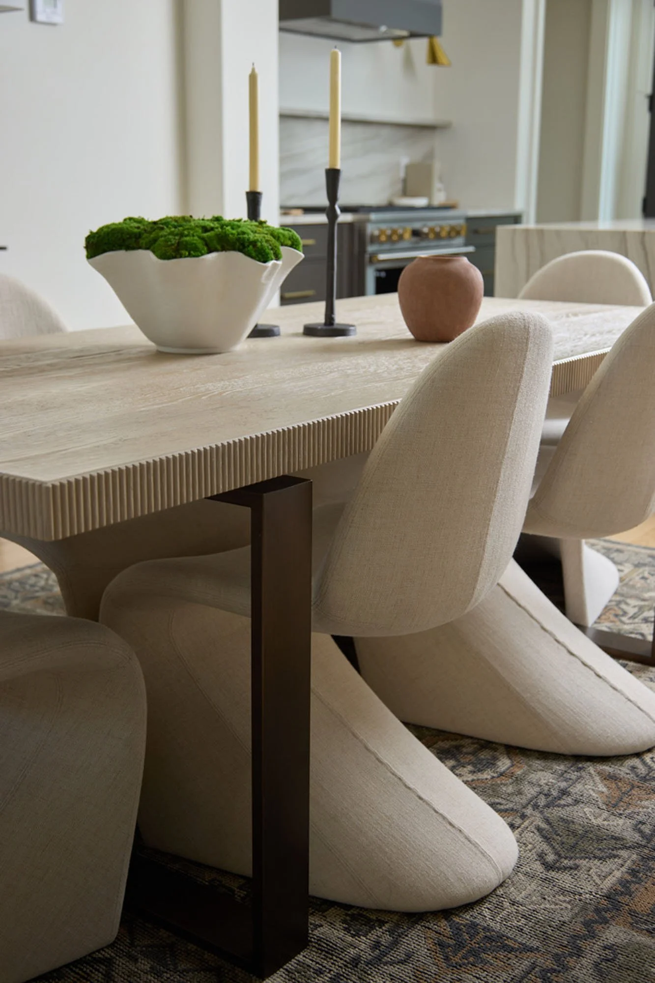Close-up of a wooden dining table with beige upholstered chairs, decorative vases, and candlesticks, with a kitchen in the background.
