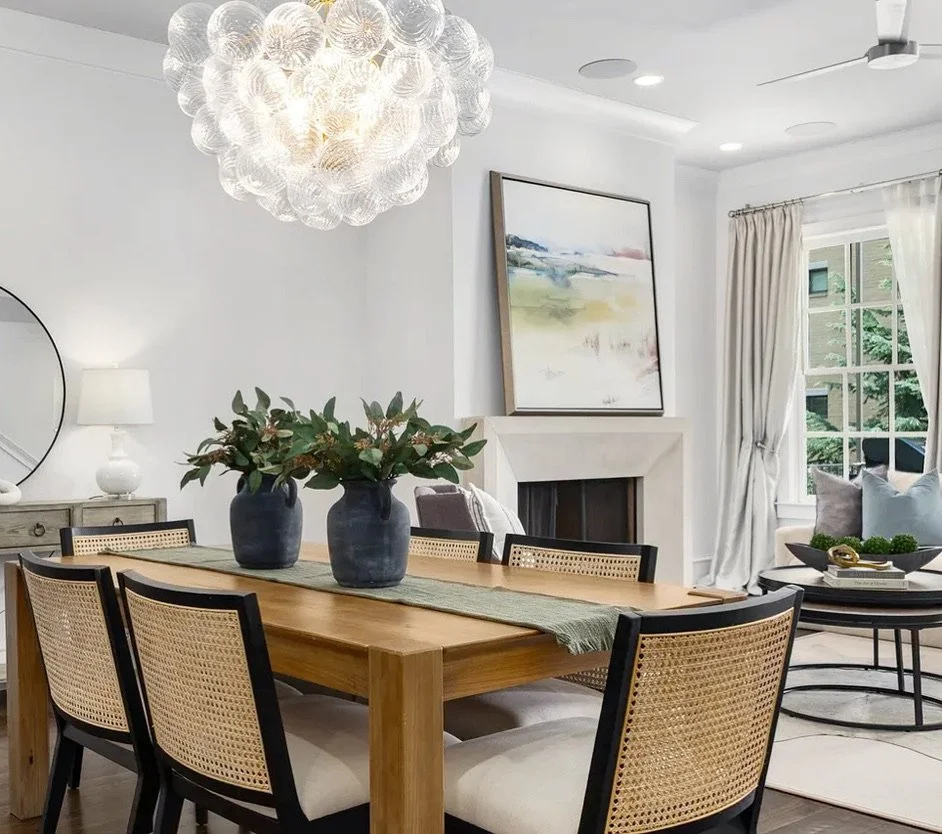 A contemporary dining and living room with a wooden dining table, six black and rattan chairs, two large vases with green foliage, a white side table with a lamp, a circular mirror, abstract painting, a white fireplace, and a large window with cream curtains.