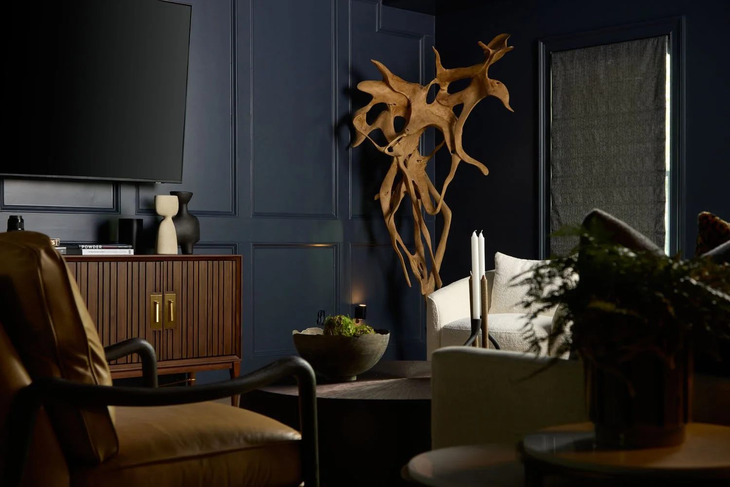 Interior of a living room with dark blue walls, a large flat-screen TV, a wooden cabinet with decorative vases, a white armchair, a modern wooden wall art piece, a black candle holder with white candles, a bowl with green plants, and a table with a plant in a dark pot.