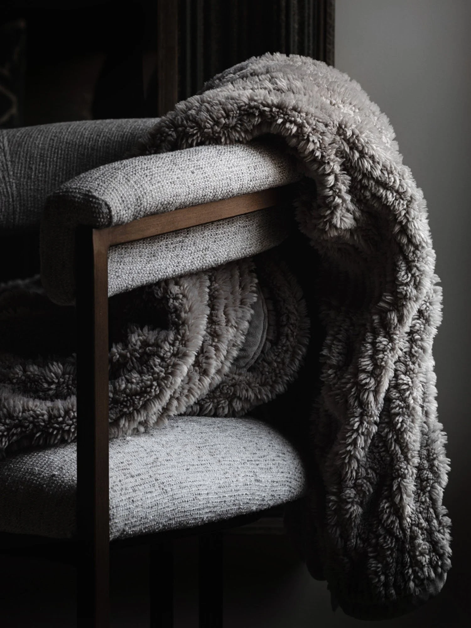 Close-up of a gray upholstered armchair with a textured fabric, topped with a thick, cozy, gray fluffy throw blanket folded over the armrest in a dimly lit room.