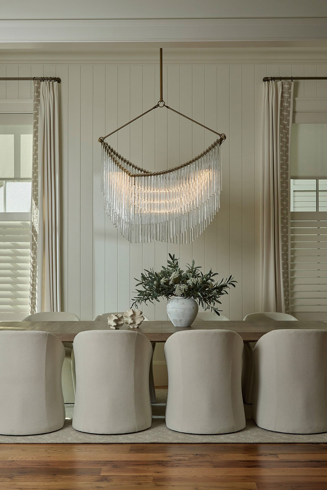 A modern dining room with a wooden table, beige upholstered chairs, and a large vase with greenery and white flowers. A chandelier hangs above the table, and windows with white curtains flank the walls.