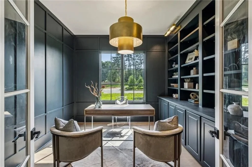 A small, modern home office or study room with dark blue walls, built-in shelves, a large window showing a green outdoor view, a rectangular wooden desk, two beige curved armchairs with cushions, and a gold pendant light hanging from the ceiling.