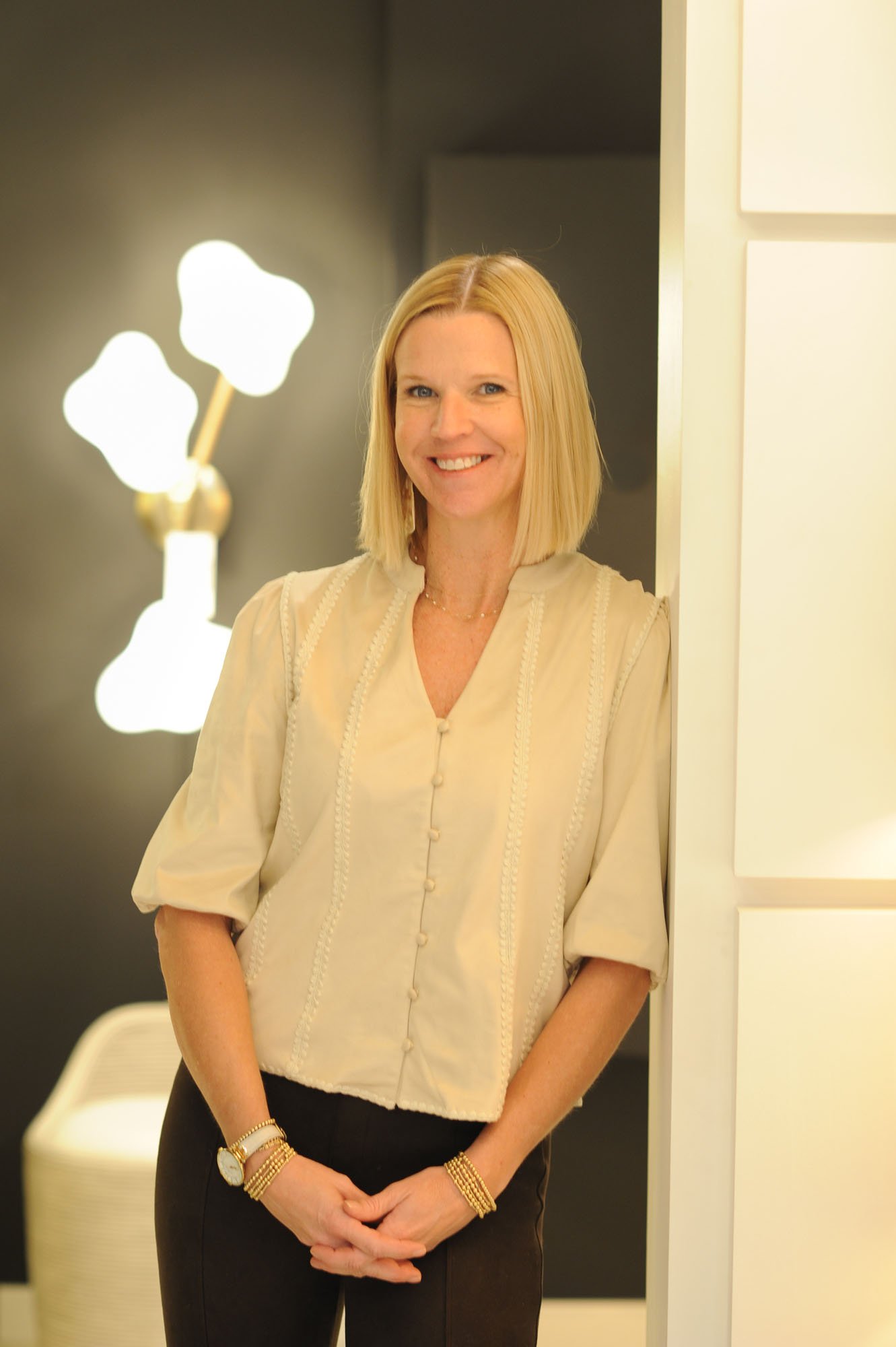 A woman with blonde hair wearing a beige blouse and black pants, standing indoors, smiling at the camera.