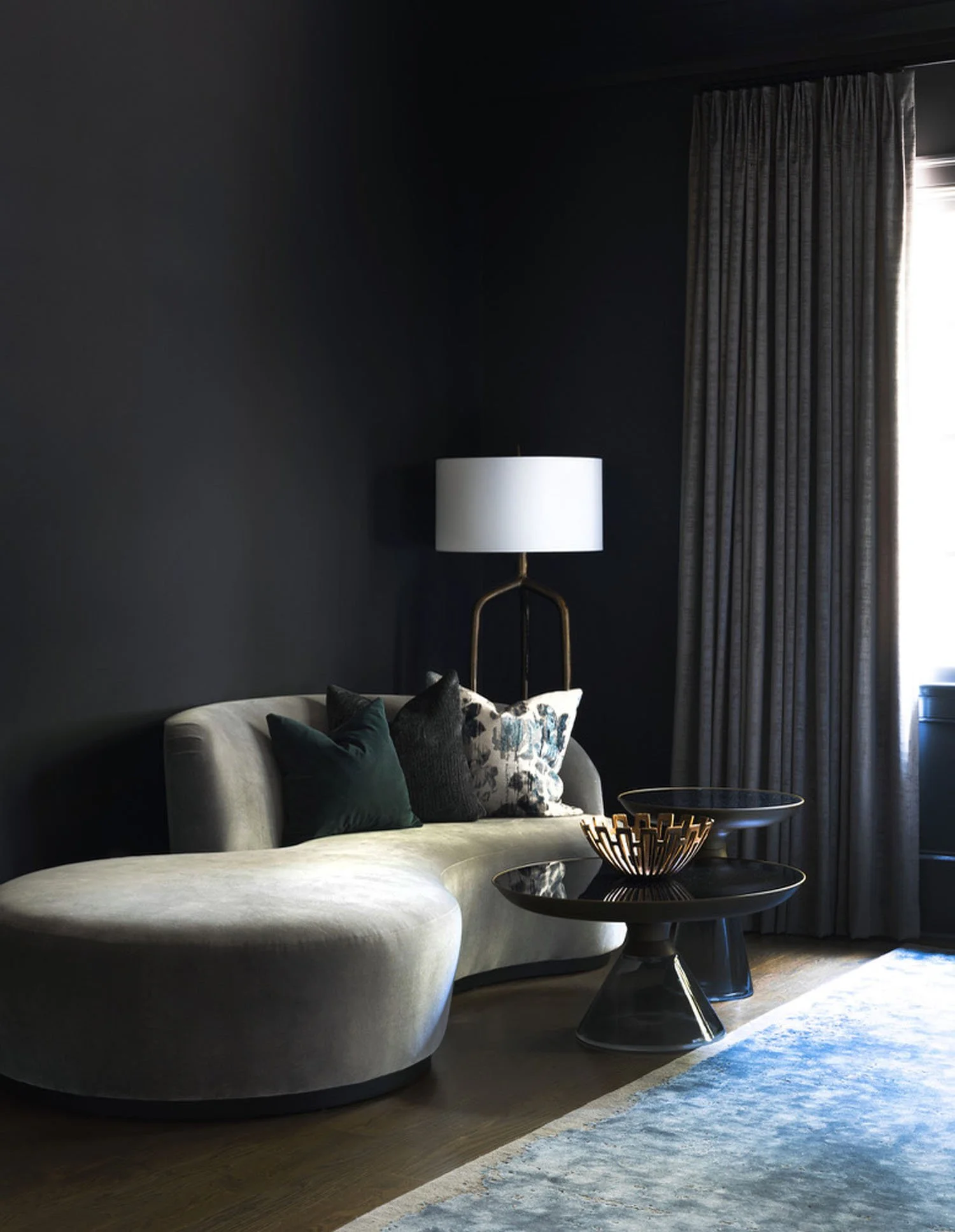 A modern living room with dark walls, a curved beige sofa with multiple decorative pillows, a black side table, and a gold decorative bowl. There are also tall curtains covering a window, and a tall white lamp with a gold stand beside the sofa.
