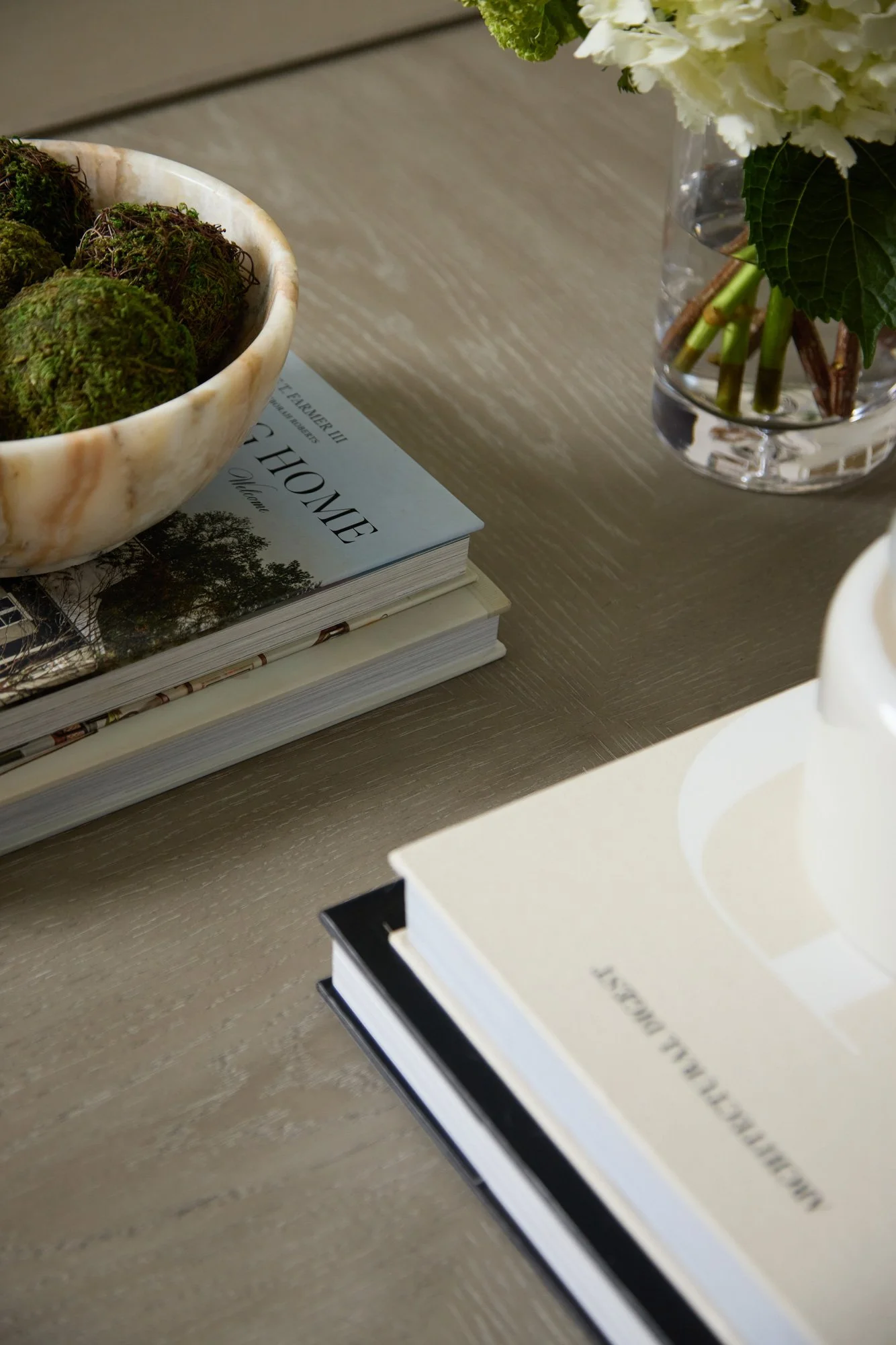 A marble bowl containing moss balls placed on top of stacked books, including one titled 'Home'. To the right, a glass vase with white flowers and green leaves is on a wooden surface.