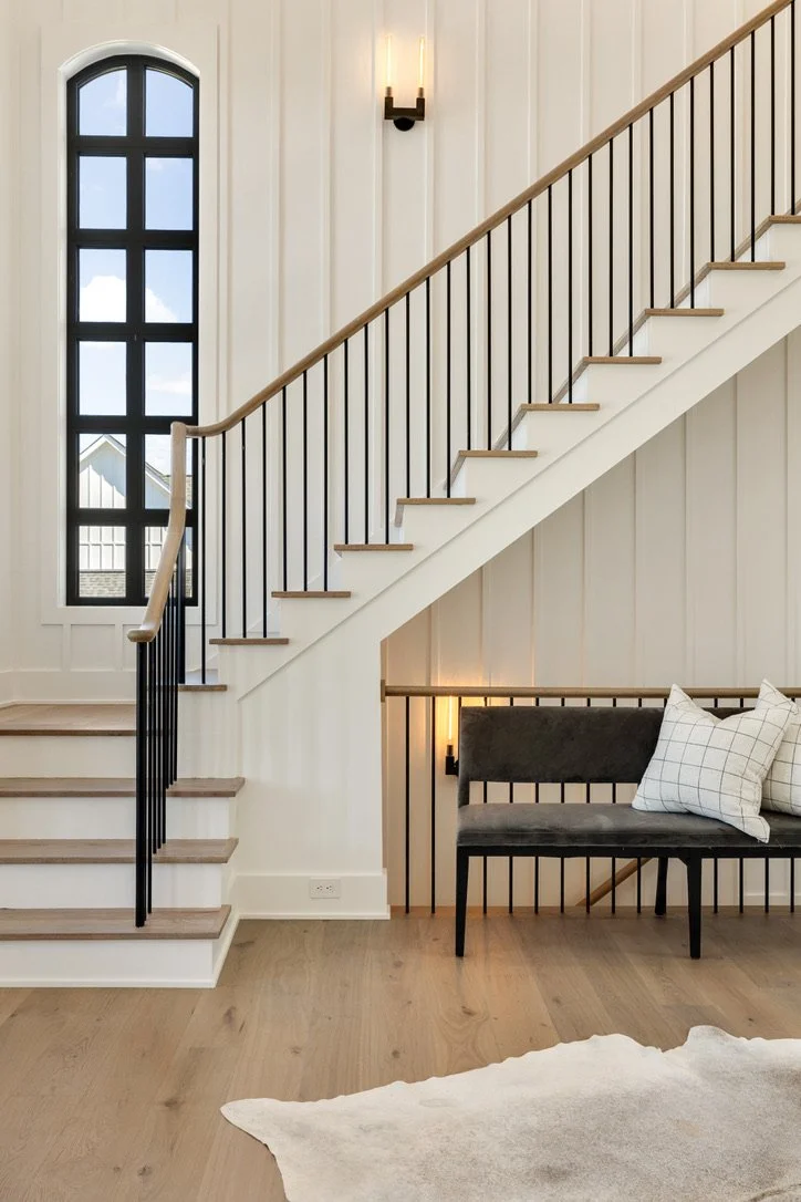 Interior view of a modern staircase with black balusters, wooden handrail, and white walls, next to a tall arched window with black framing, and a black bench with pillows below the stairs. There is a wall sconce light and a light-colored wooden floor.