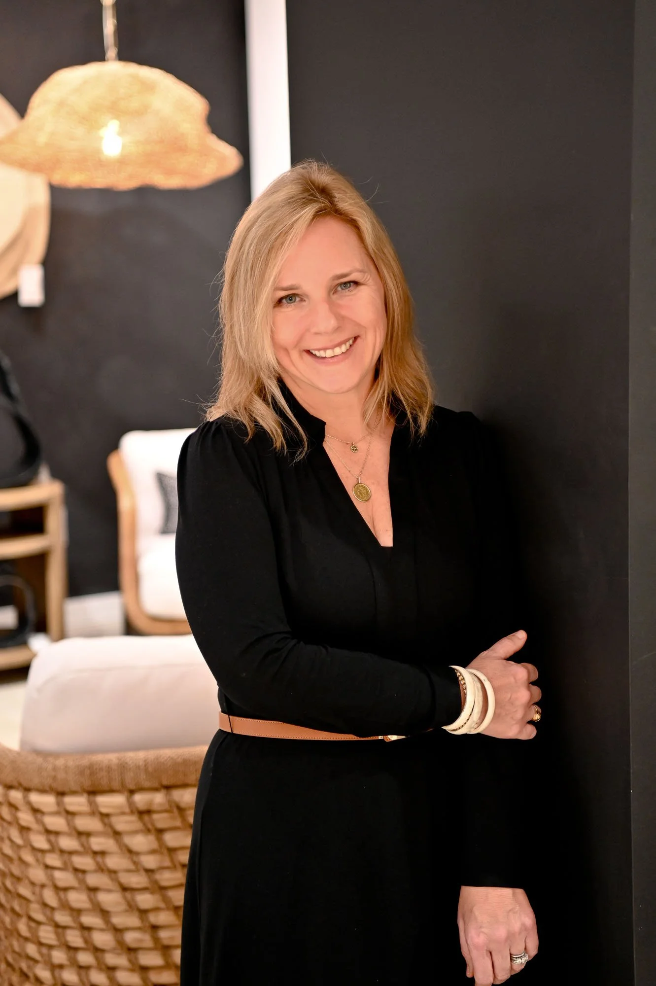 A smiling blonde woman wearing a black dress, standing in a room with modern decor, including a hanging lamp and a woven chair.
