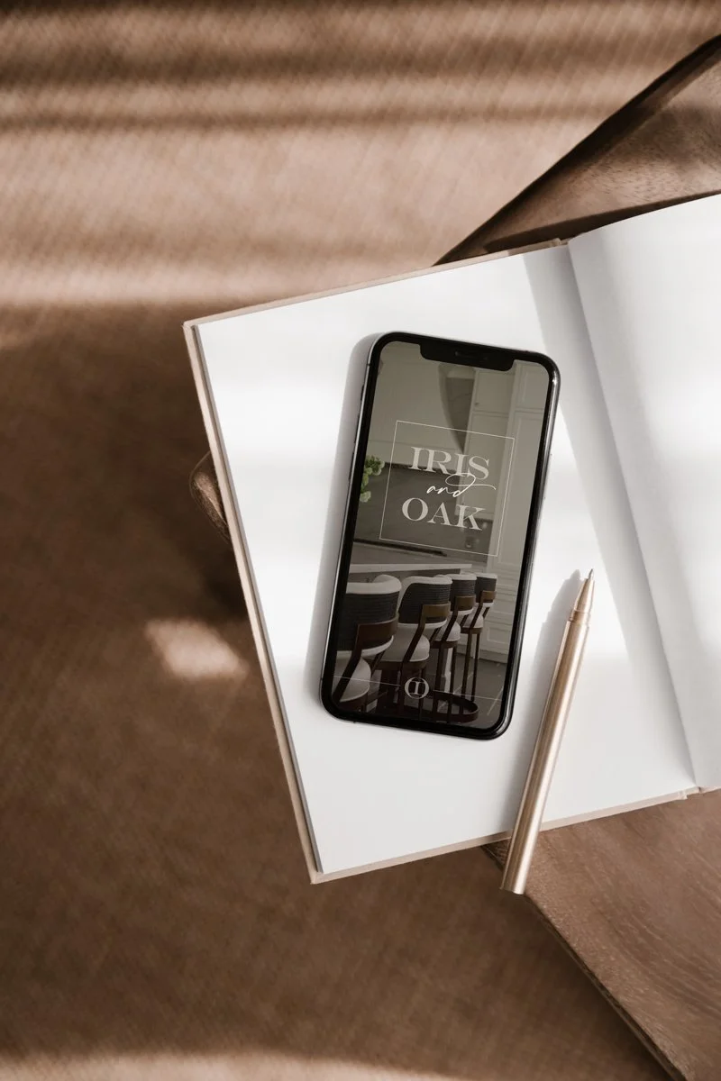 A smartphone displaying the text 'IRIS and OAK' resting on an open white notebook, with a cream-colored pen beside it, on a wooden table.