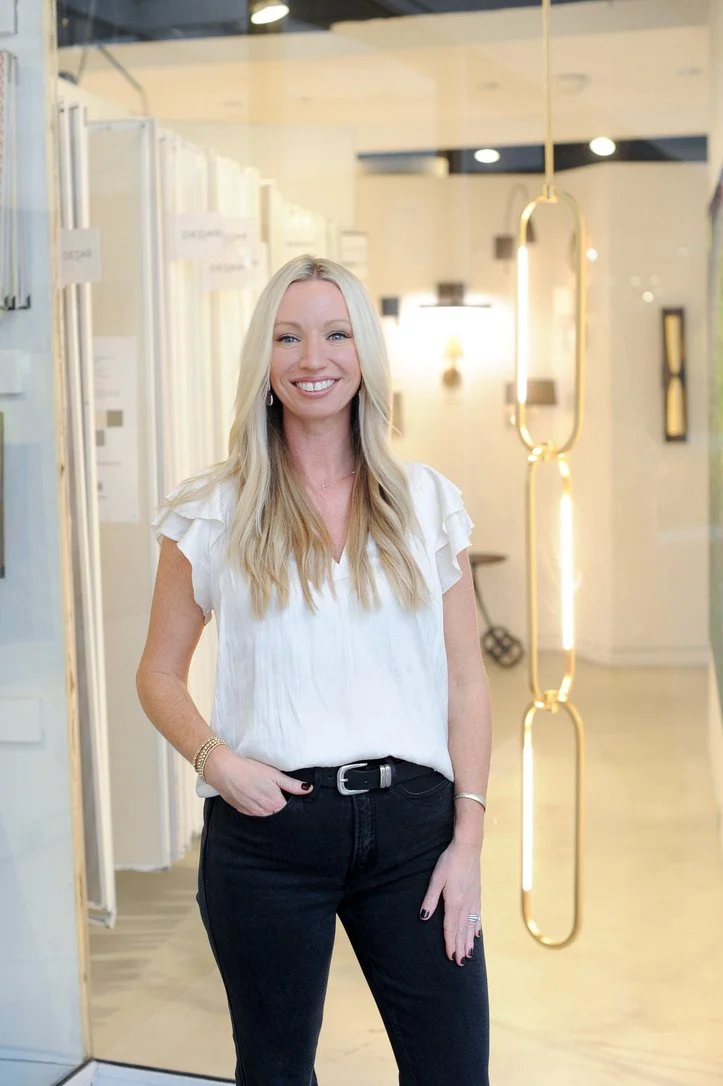 A smiling woman with long blonde hair wearing a white blouse and black pants standing inside a modern, well-lit interior space.