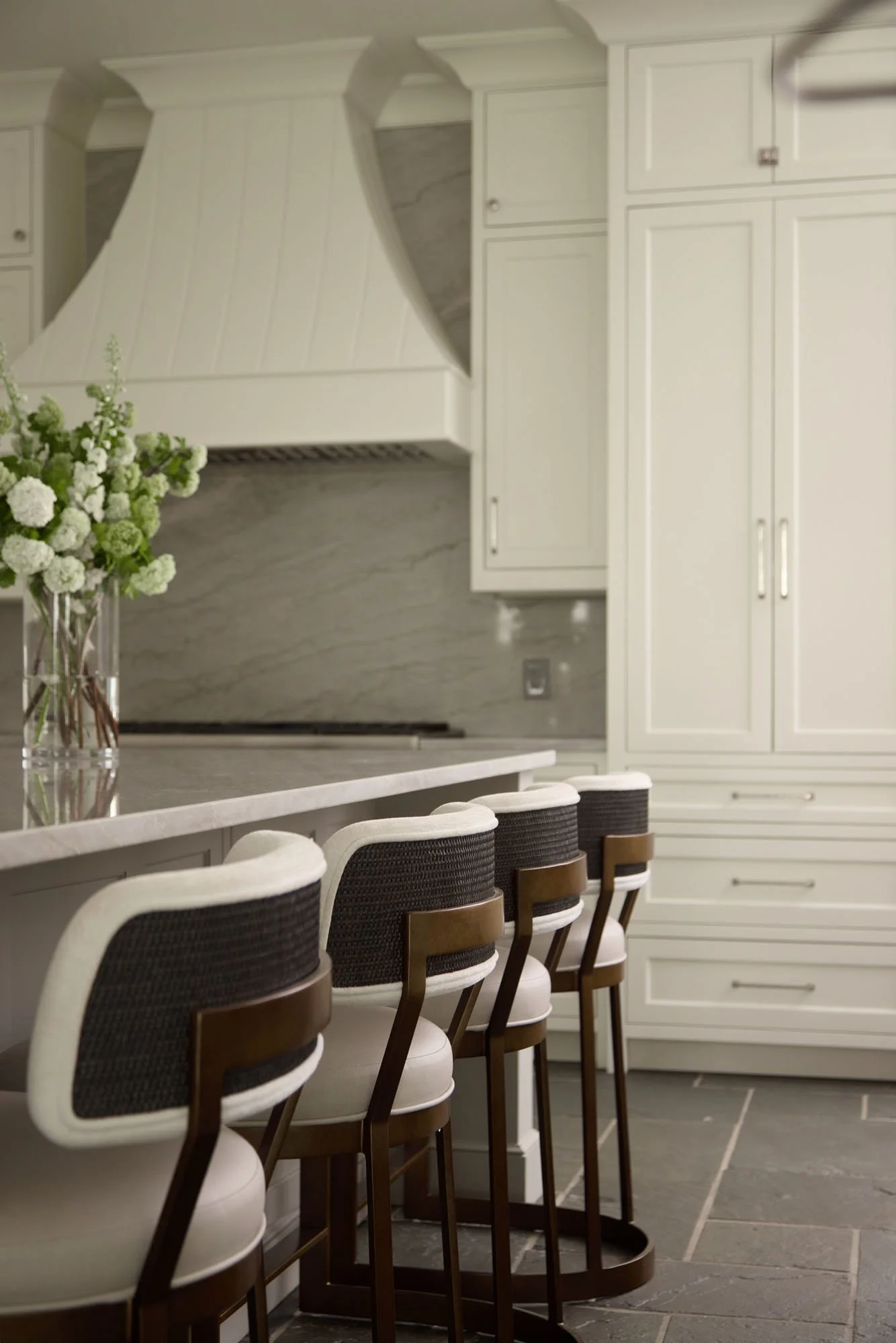 Kitchen with white cabinets, dark bar stools with white seats, and a vase of white and green flowers on the marble counter.