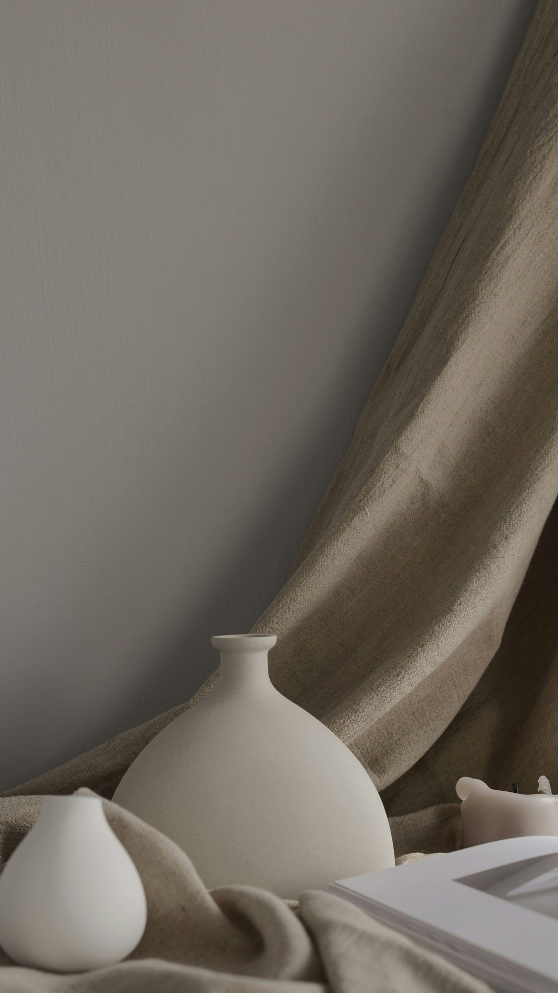 Close-up of a white ceramic vase on a beige cloth, with a beige curtain and plain wall in the background.