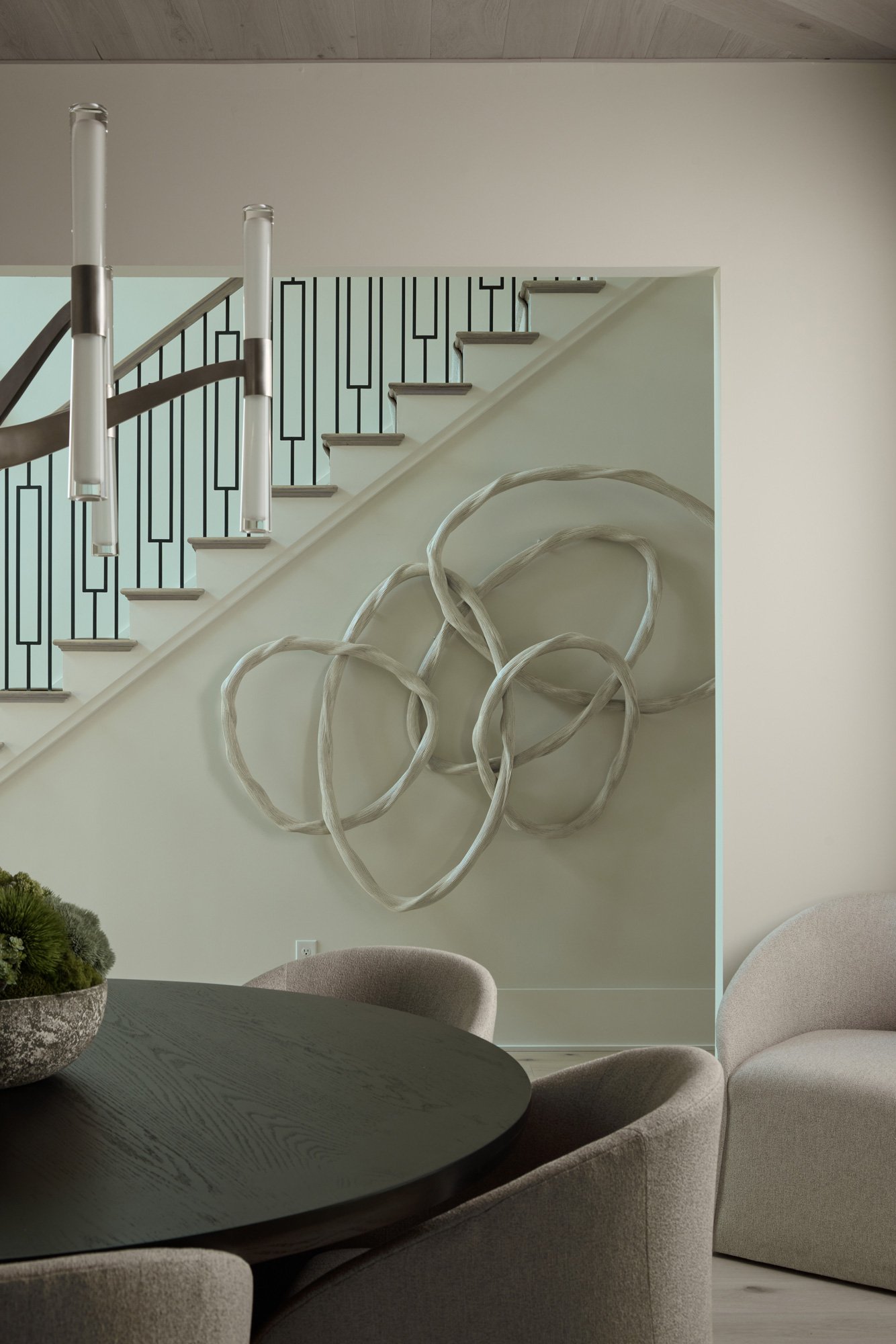 Interior of a modern home with a staircase featuring artistic metal railings, a dark wooden table with beige upholstered chairs, a decorative plant on the table, and a wall art piece with intertwined loops.