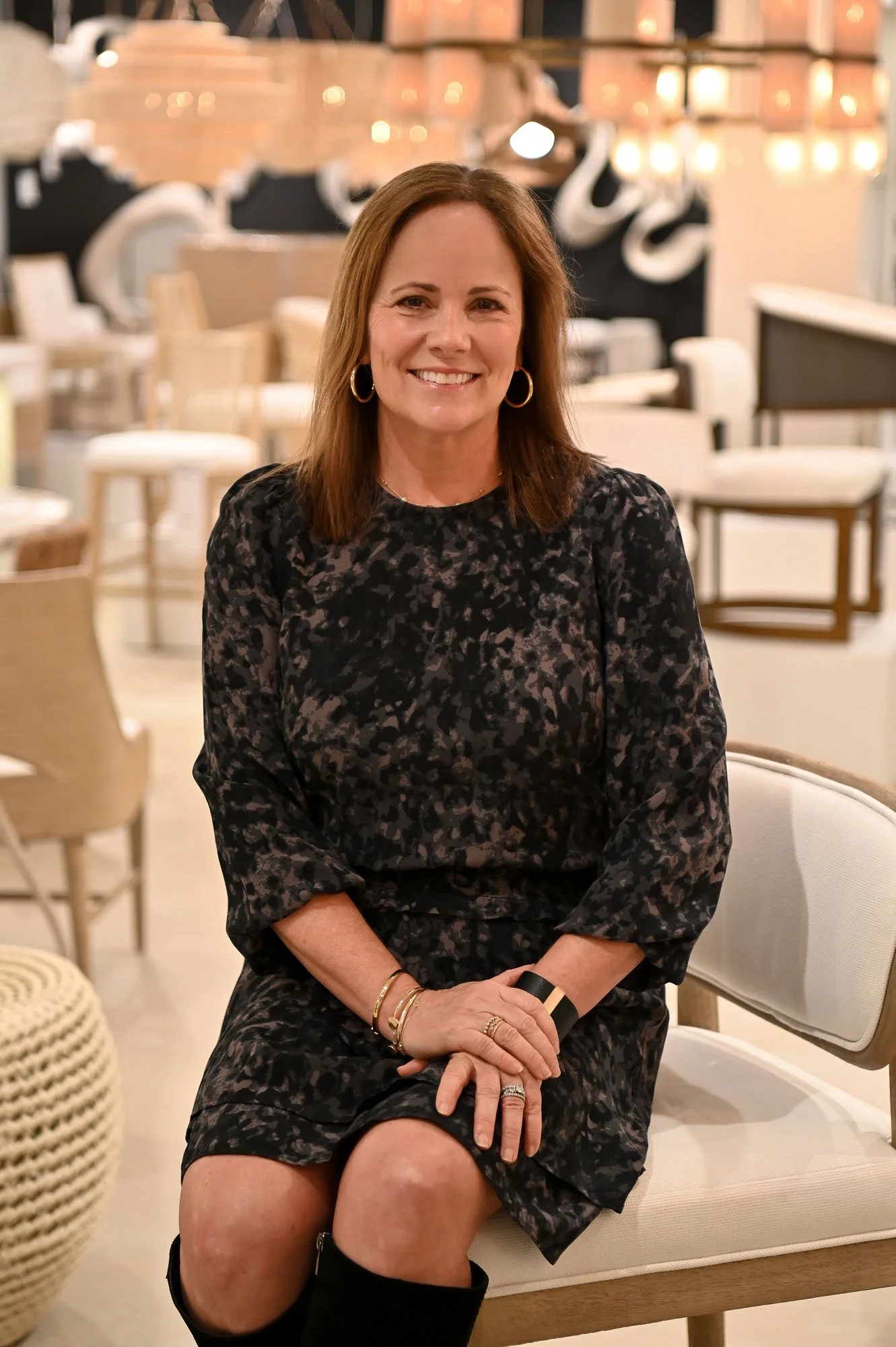 A woman with light skin, brown hair, and hoop earrings, smiling, sitting on a beige chair in a well-lit, modern cafe or restaurant with white chairs, tables, and hanging lights.