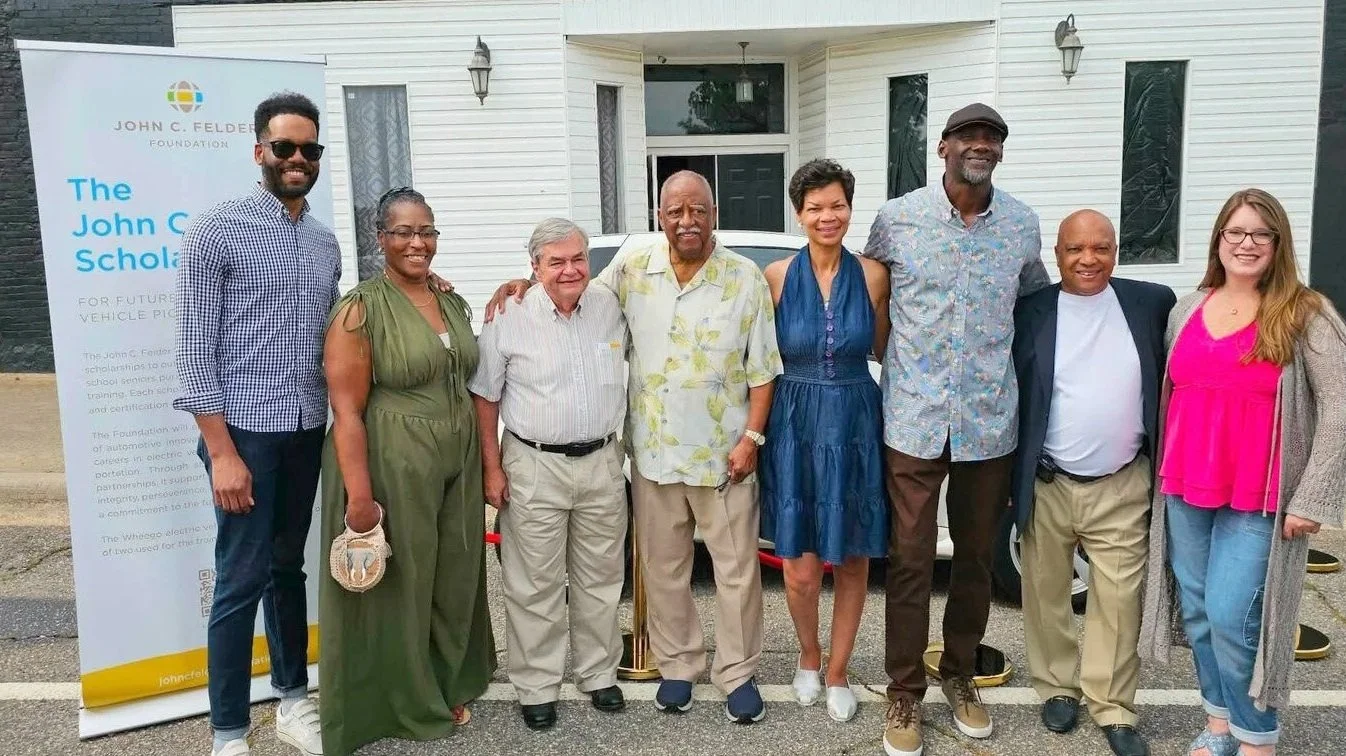A group of nine diverse people standing outdoors in front of a white house and a banner for The John C. Felder Foundation, smiling for a photo.