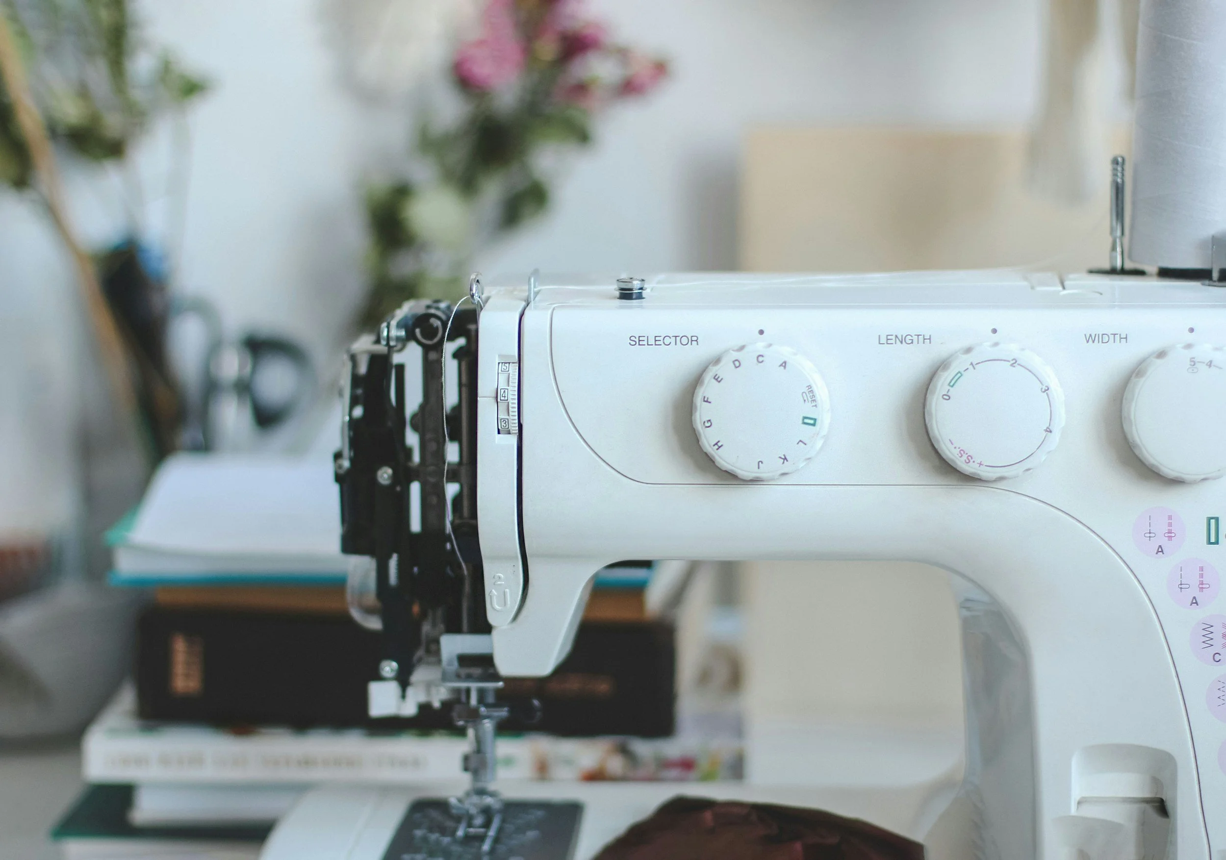 Close-up of a white sewing machine with dials labeled 'SELECTOR', 'LENGTH', and 'WIDTH' in focus, with blurred sewing area and spools of thread in the background.