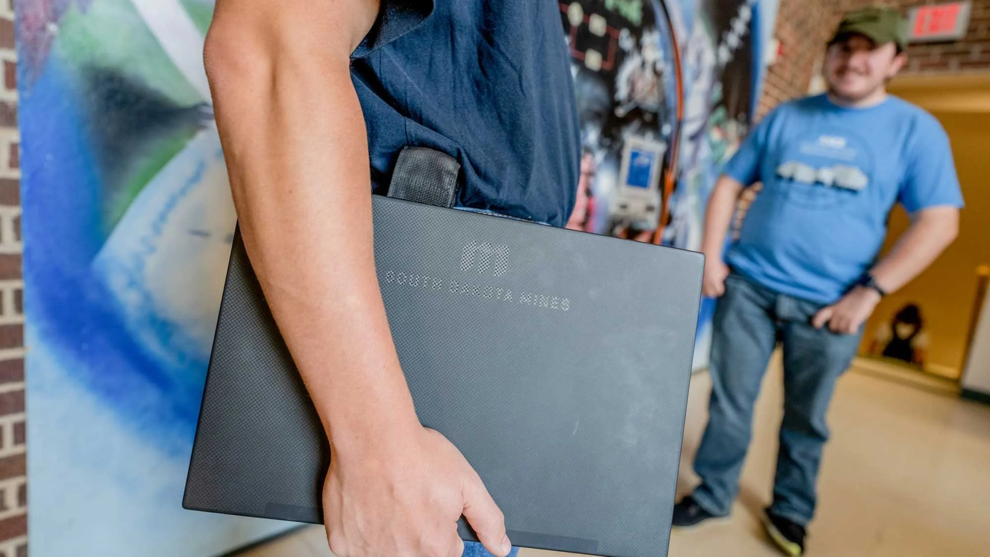 Person holding a laptop with South Dakota Mines text, standing next to a mural, with a man in a blue shirt and cap in the background.