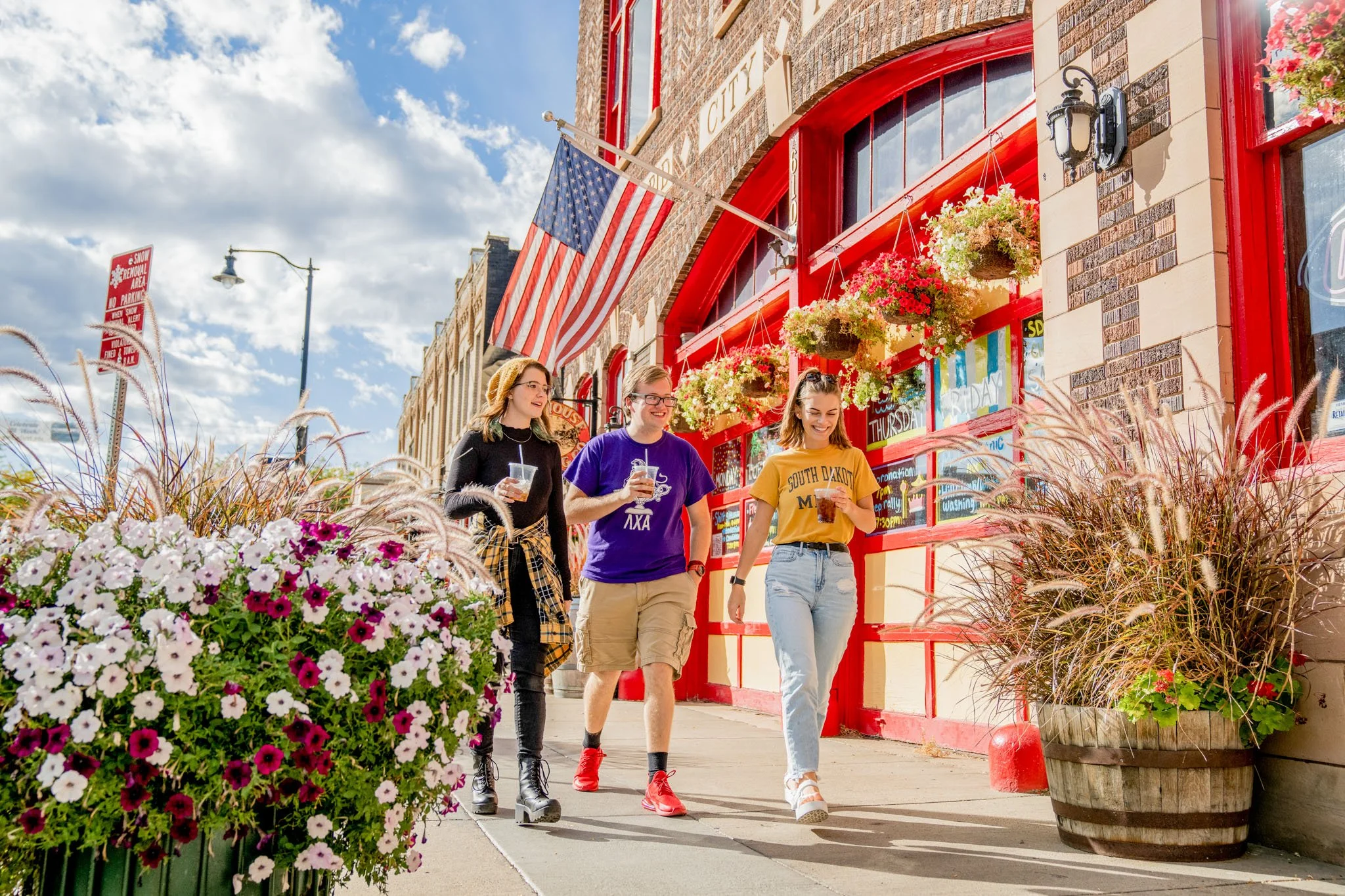 Three young women walking and smiling on a city sidewalk, with a red building decorated with hanging flower baskets and an American flag in the background.