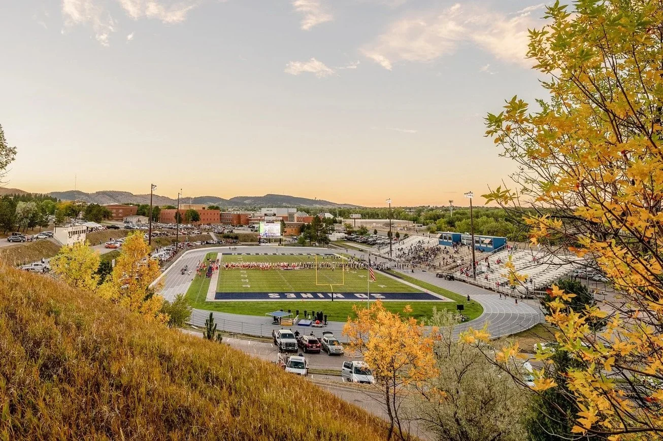 A football stadium during sunset with cheerleaders in formation on the field, surrounded by a running track, parking lots, trees with fall foliage, and distant hills.