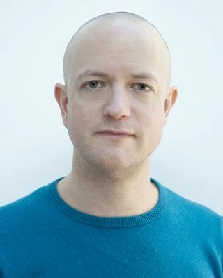 Portrait of a bald man with light skin, blue eyes, and a slight smile, wearing a blue sweater against a plain light background.