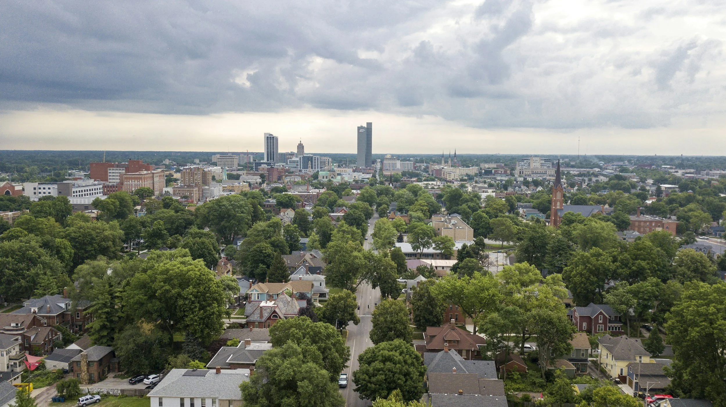 A cityscape with a mix of residential houses and larger commercial buildings under an overcast sky, with trees lining the streets and the downtown skyline visible in the distance.