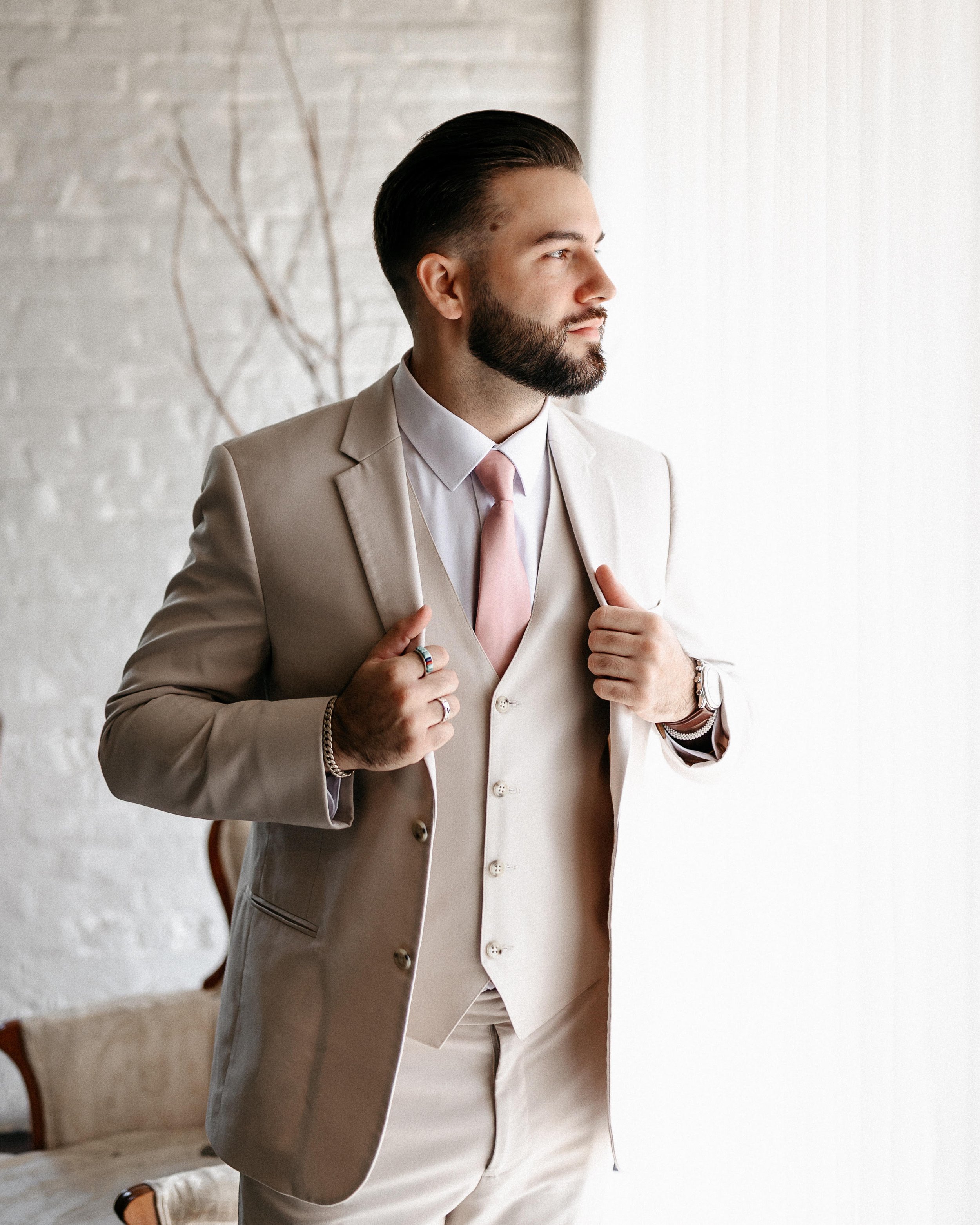 A man with a well-groomed beard and slicked-back hair is wearing a beige suit, white shirt, and light pink tie. He is adjusting his suit jacket with both hands. The background is a minimalistic white stone wall with some plant branches visible.