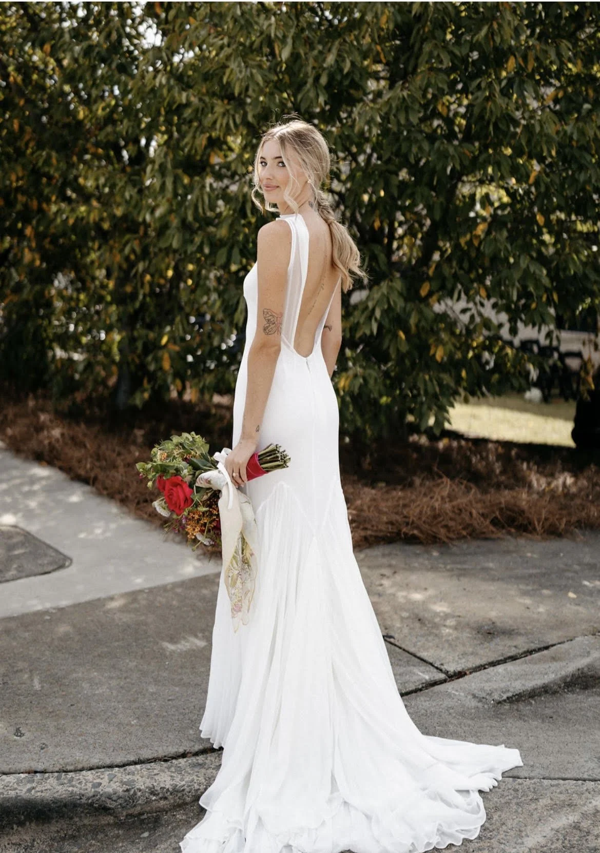 A woman in a white wedding dress holding a bouquet of red and white flowers standing outdoors near green trees.