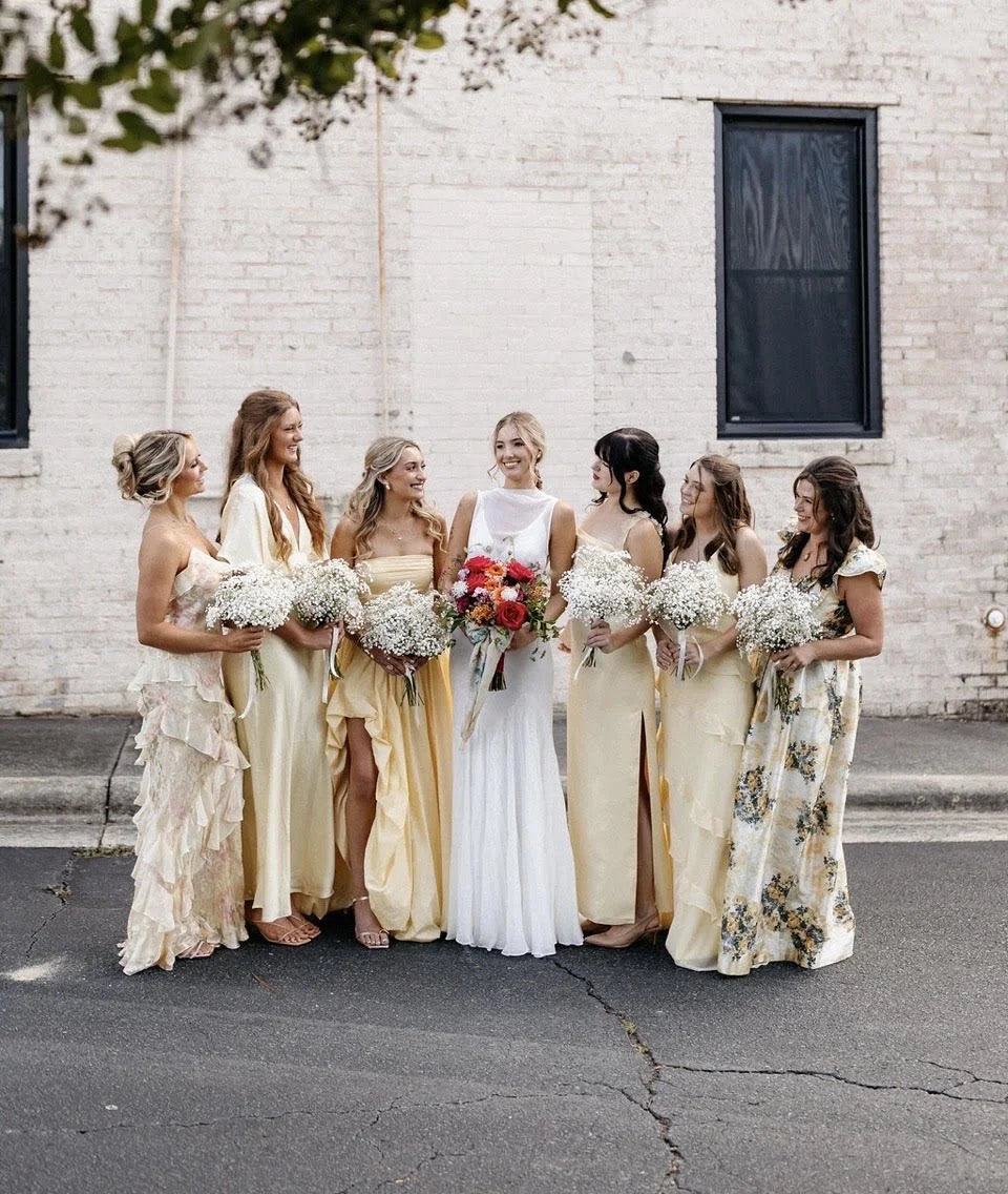 A group of seven women in dresses standing on a city street, holding bouquets of flowers, with a white brick wall in the background.