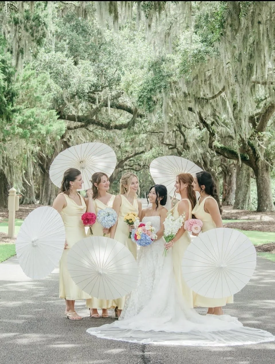 A bride and five women in matching yellow dresses holding bouquets of colorful flowers in a park with large oak trees, white parasols, and a pathway.