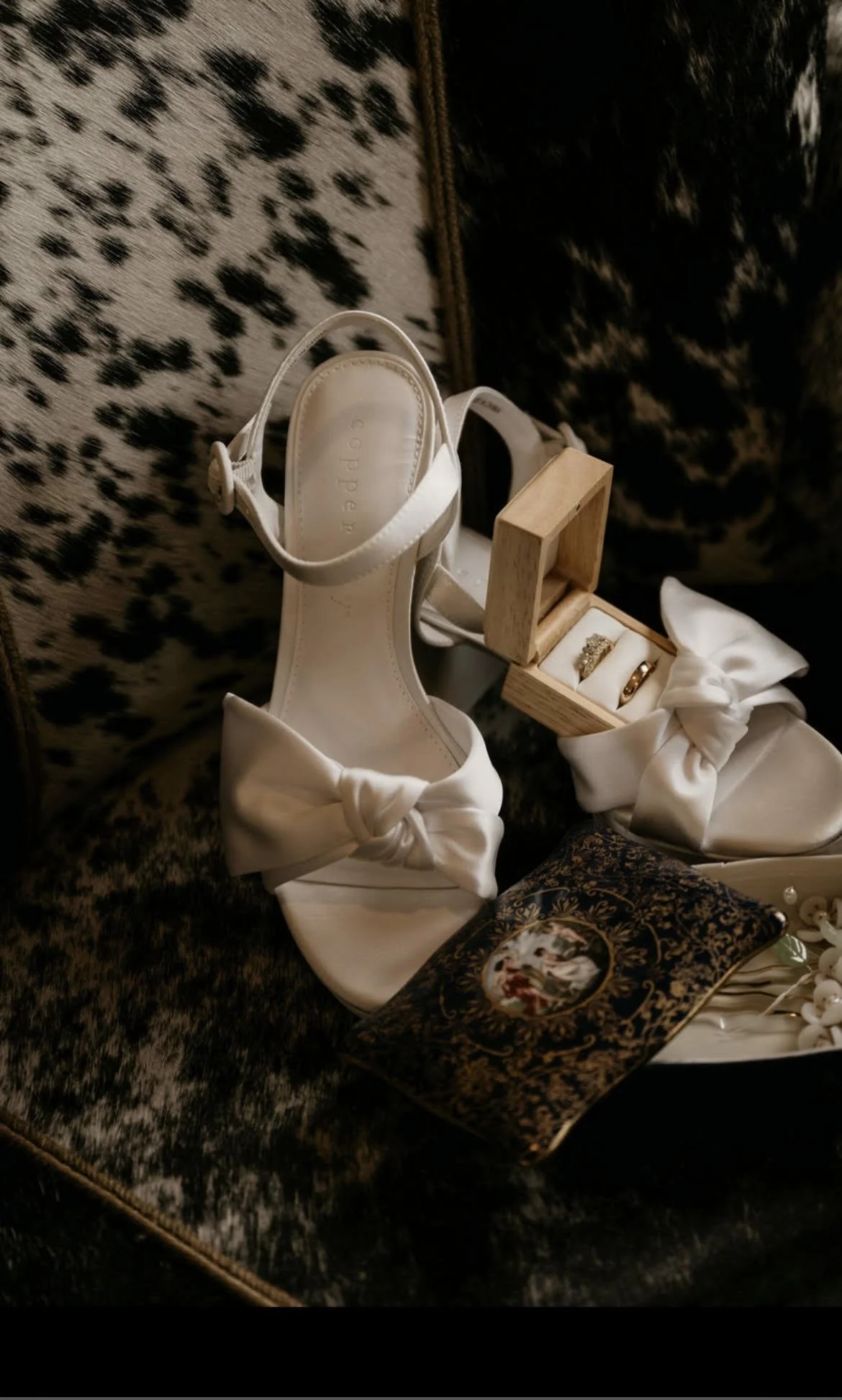 A pair of white bridal shoes with bows, a small wooden box with rings, a decorative pillow, and a floral wedding booklet on a leopard print fabric surface.