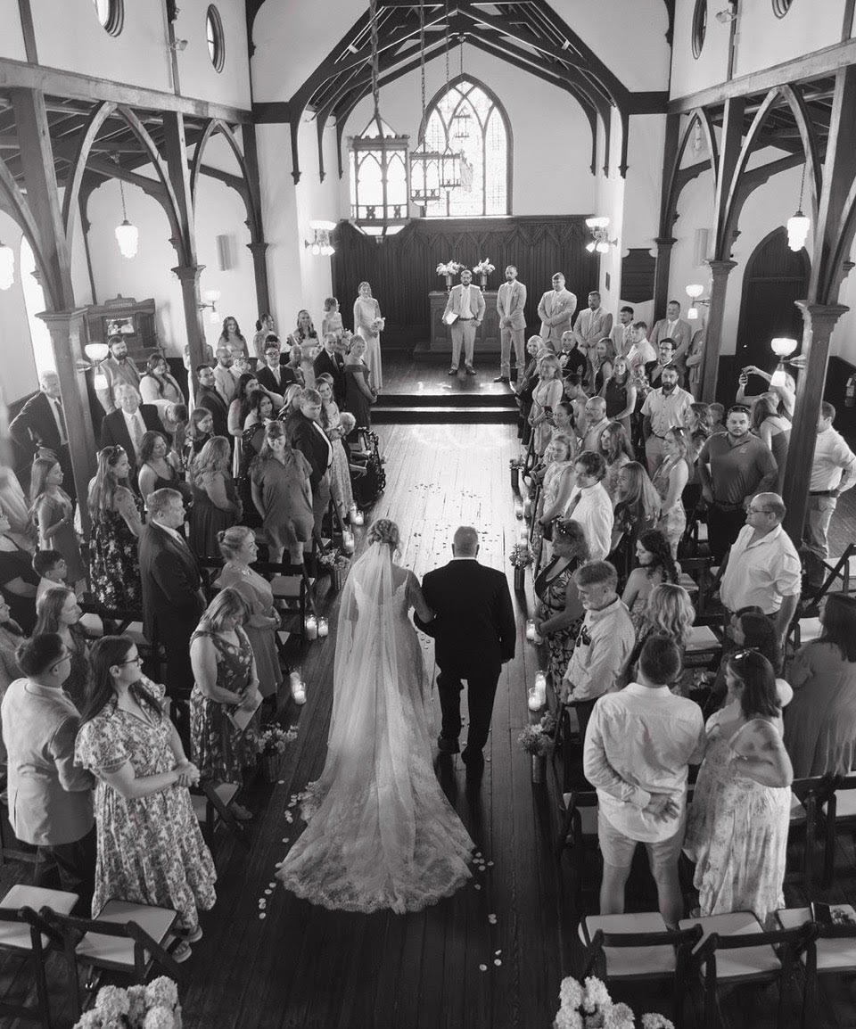 Black and white photo of a wedding ceremony inside a church. The bride, dressed in a long gown with a train, is walking down the aisle with a man, likely the groom. Guests are seated and standing on either side of the aisle, watching the procession. The church has high arched ceilings, stained glass windows, and wooden beams.