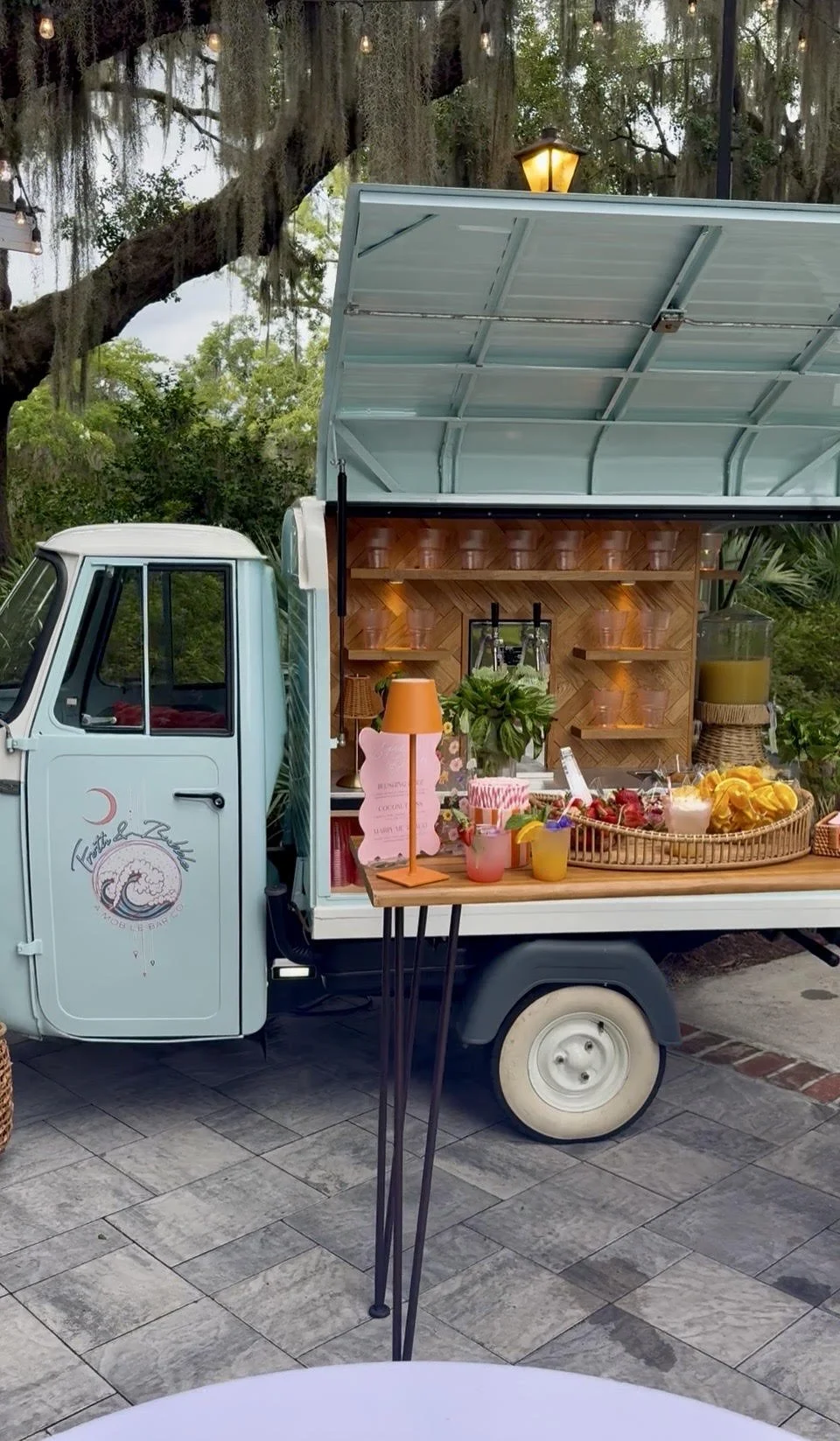 A vintage light blue food truck with open roof and serving window, decorated with a tropical theme, featuring a small table with drinks and a pink lamp, set outdoors with trees and Spanish moss in the background.