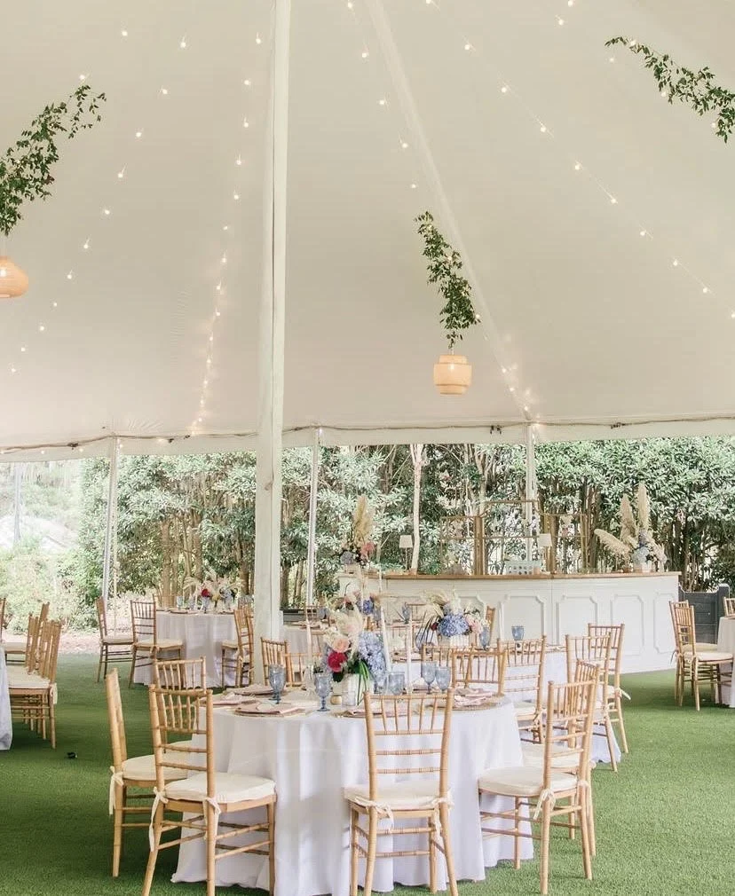 Wedding reception setup in a white tent with round tables, gold chiavari chairs, and floral centerpieces, surrounded by greenery with string lights hanging from the ceiling.