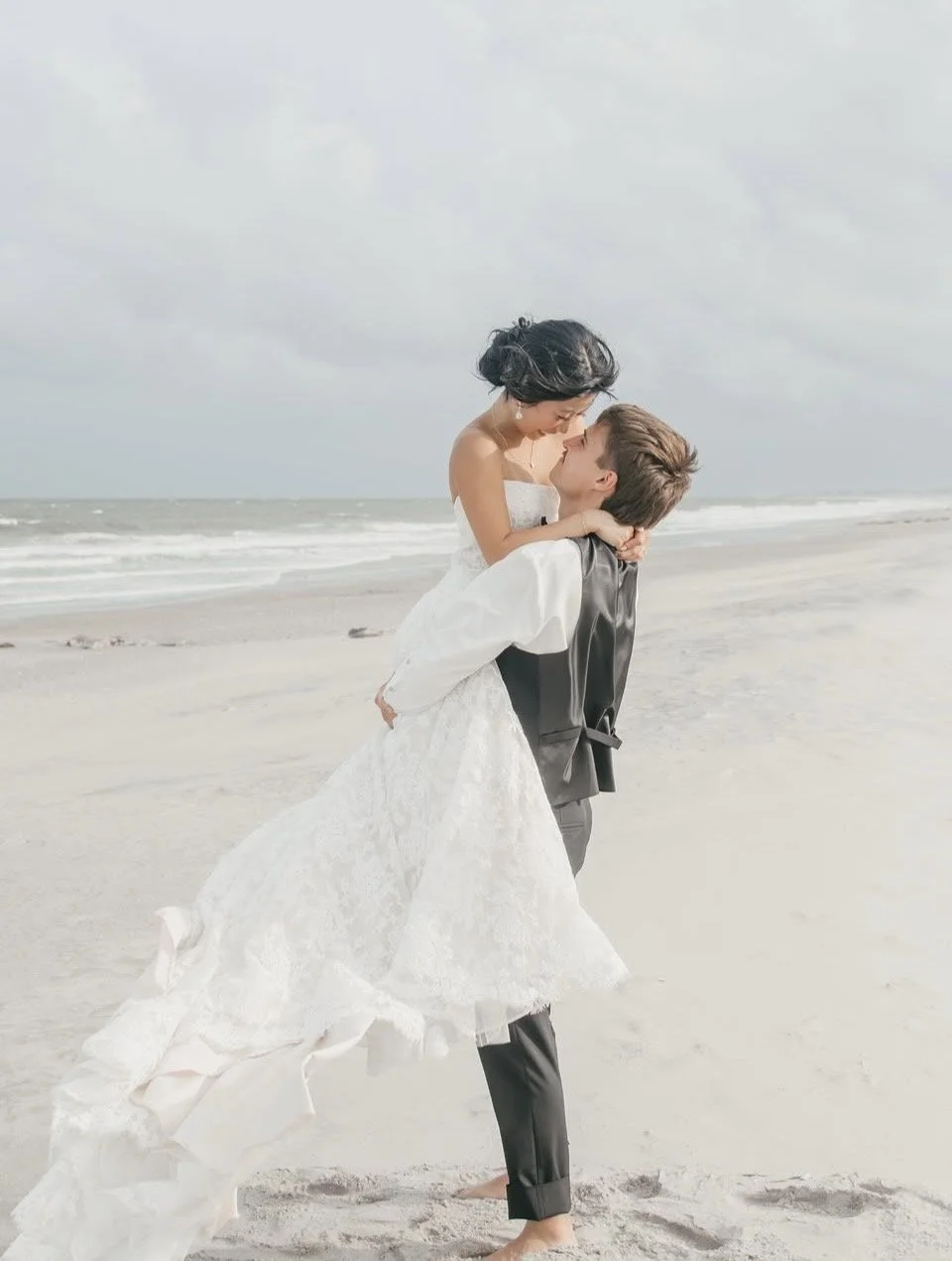 A couple dressed in wedding attire on a beach, the man lifting the woman in his arms as they look at each other.