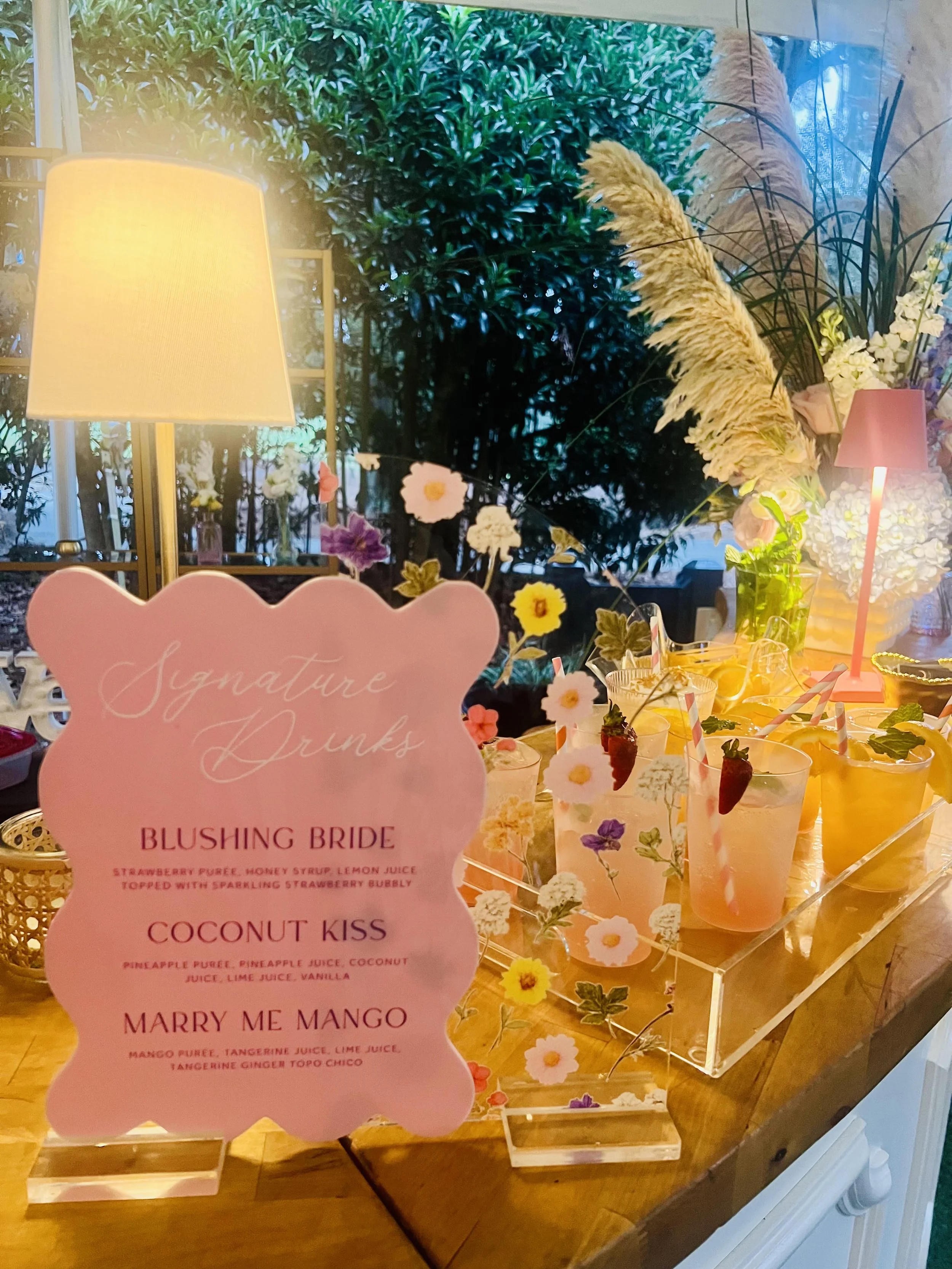 A menu card shaped like a pink bear with decorative flowers attached, displaying three drink names and ingredients: 'Blushing Bride,' 'Coconut Kiss,' and 'Marry Me Mango.' In the background, a table is decorated with colorful drinks, flower arrangements, and lamps, with greenery and plants outside visible through a window.