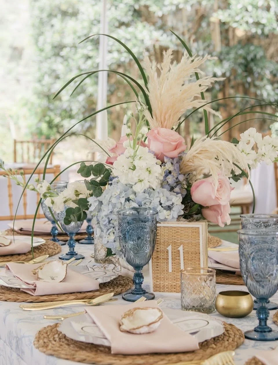 Table centerpiece with pink roses, white hydrangeas, tall grass, and pampas grass in a woven basket, set for a formal event with blue glassware, gold utensils, blush napkins, and a table number "11" in a rattan frame, outdoors with trees in the background.