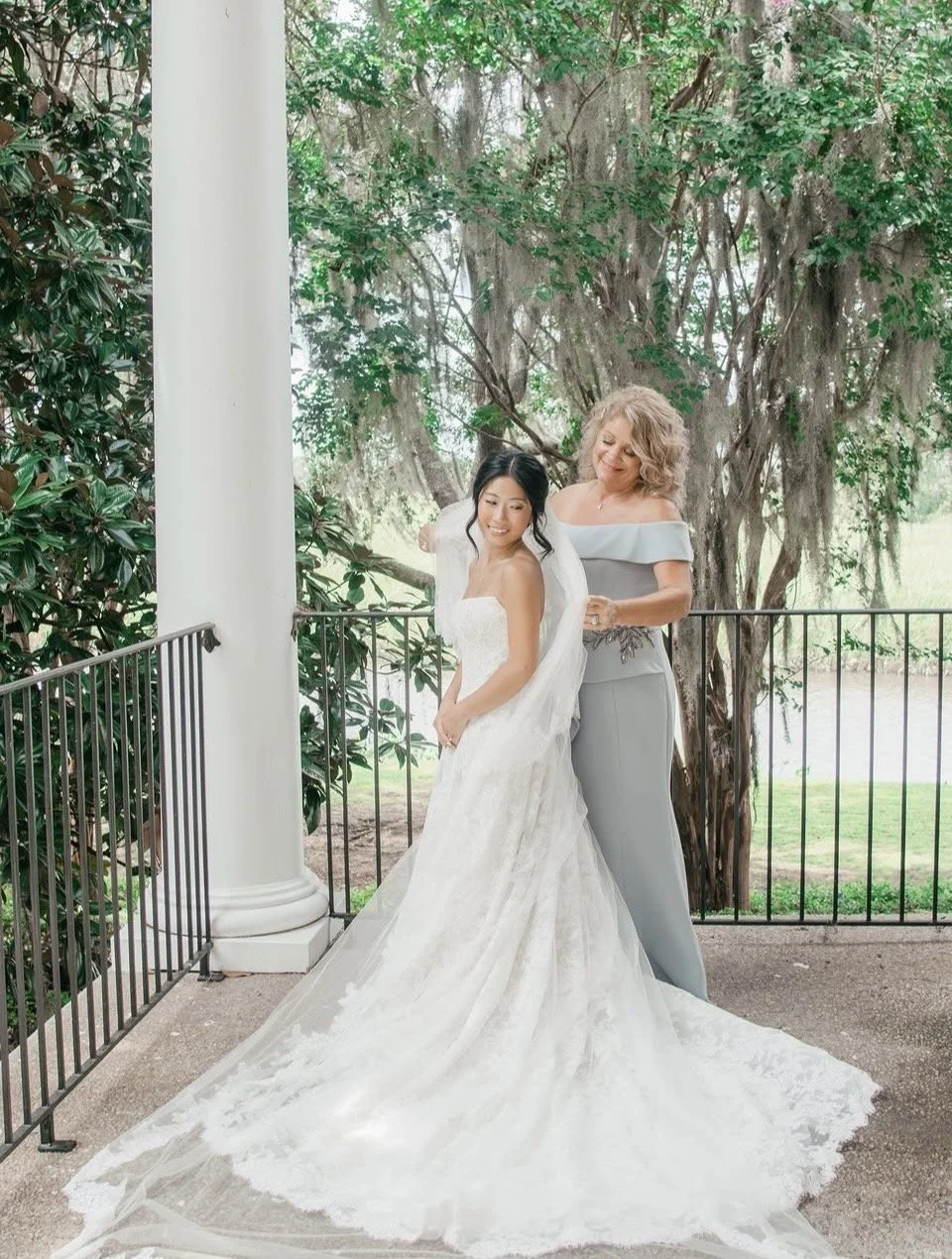 A bride in a white wedding dress with a long train standing on a porch with greenery, receiving help from a woman in a gray off-shoulder dress.