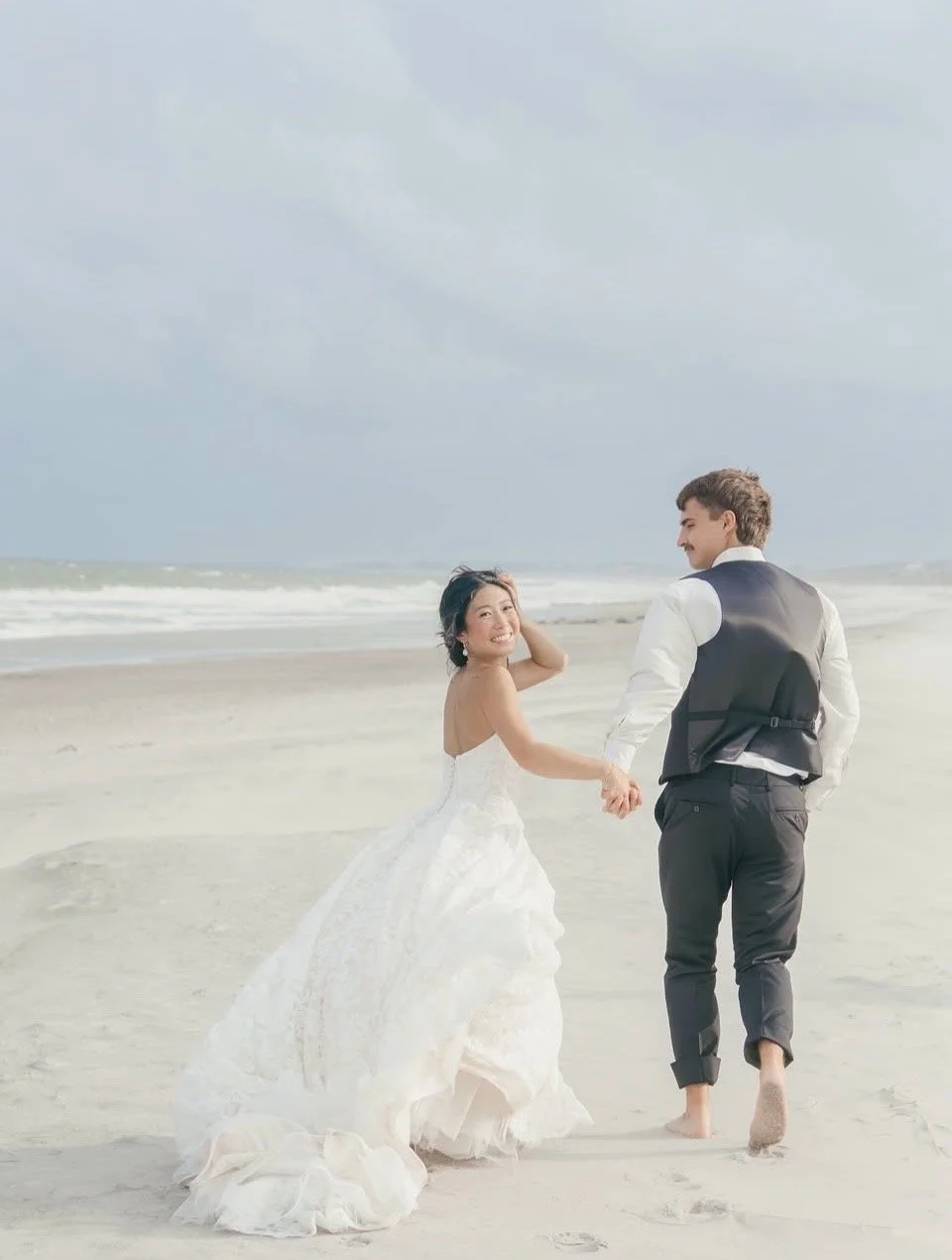A bride and groom holding hands and walking barefoot on a beach, smiling at each other with ocean waves in the background.