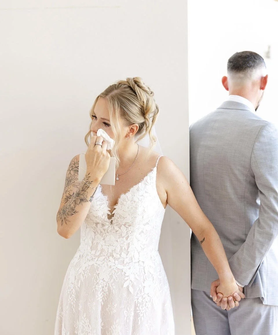 Bride in a lace wedding dress crying and wiping her tears while holding hands with her groom, who is wearing a light gray suit, backstage or at a wedding venue.
