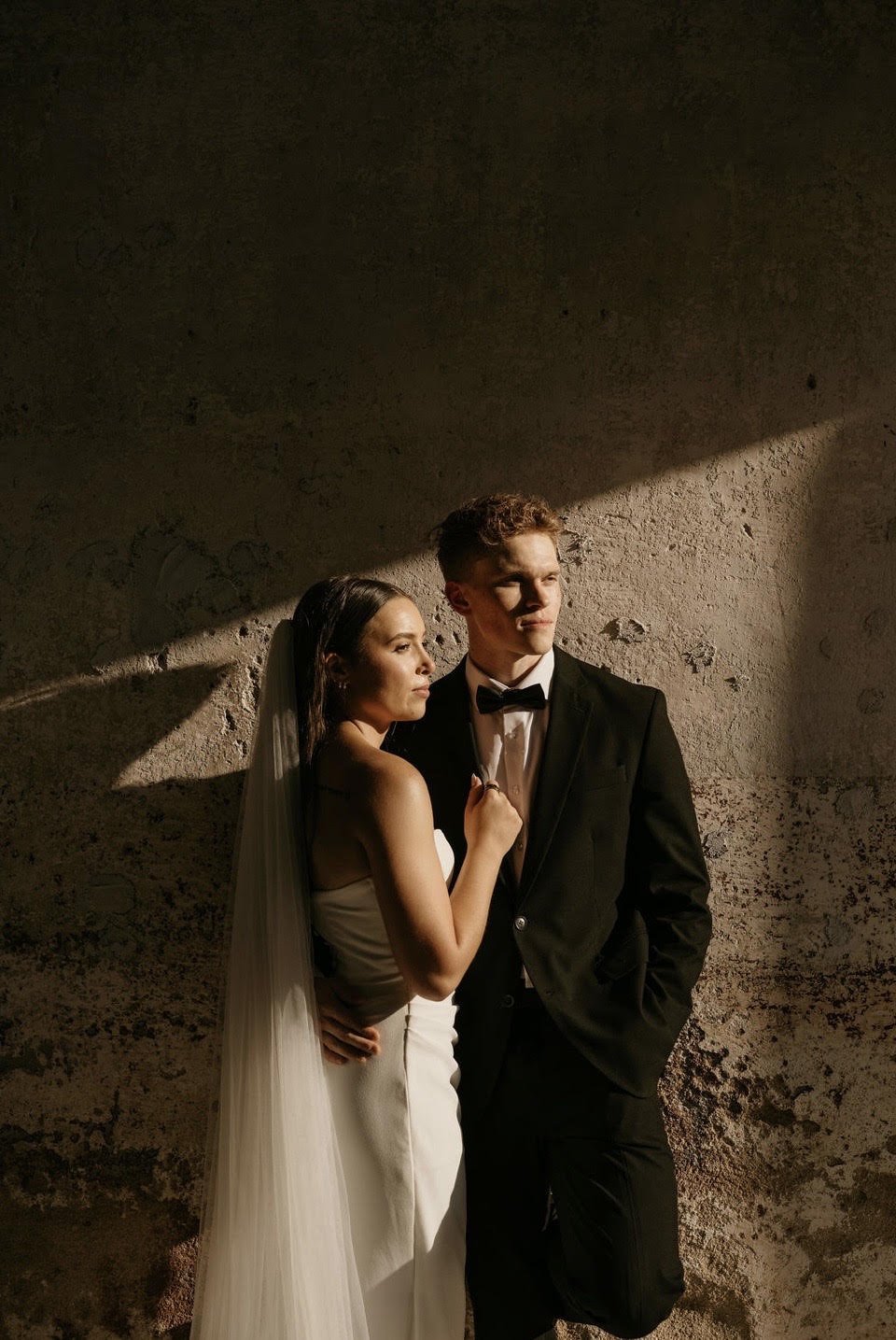 A couple dressed in wedding attire posing against a textured wall with dramatic lighting.