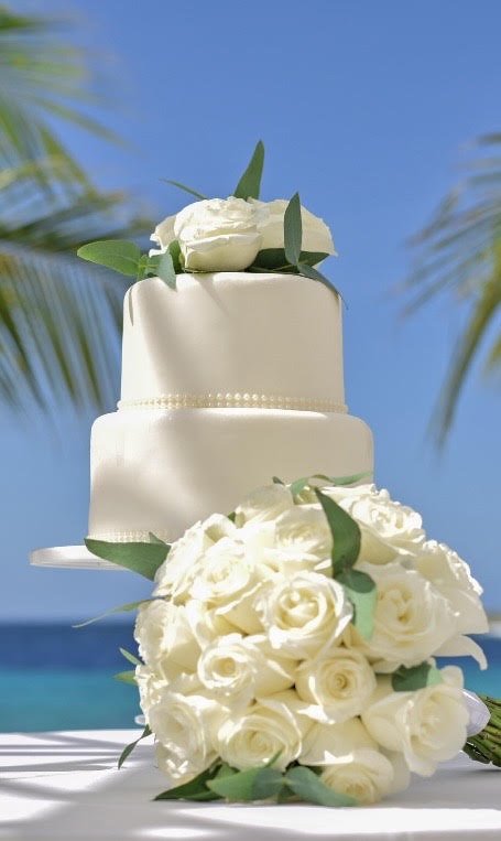 A wedding cake decorated with white roses and green leaves, set against a beach background with blue sky and palm trees.