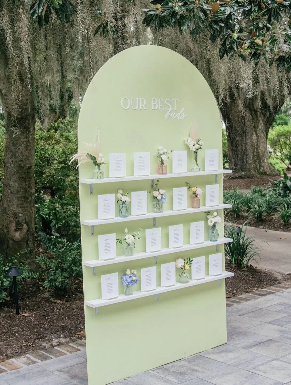 A worded wedding seating chart displays table cards and flower vases on a green wall with white shelves, located outdoors among trees and greenery.