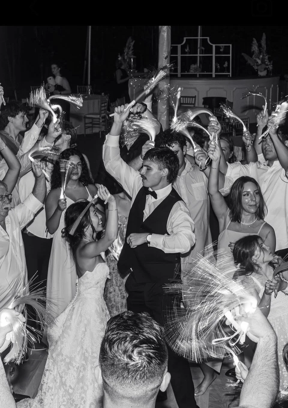 A black and white photo of a lively celebration with people dancing, holding sparklers and party horns, including a man in a tuxedo and a woman in a white dress, at what appears to be a wedding reception or festive event.