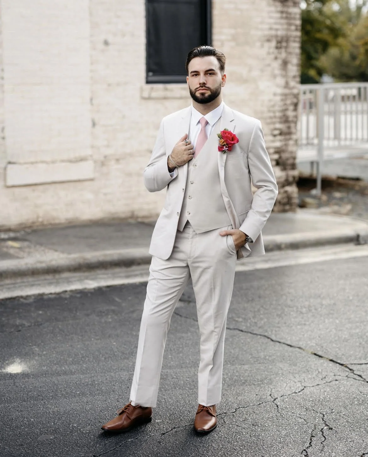 A man in a white suit with a pink tie and a pink flower boutonniere, standing outdoors on a street, with a serious expression and his hand adjusting his tie.