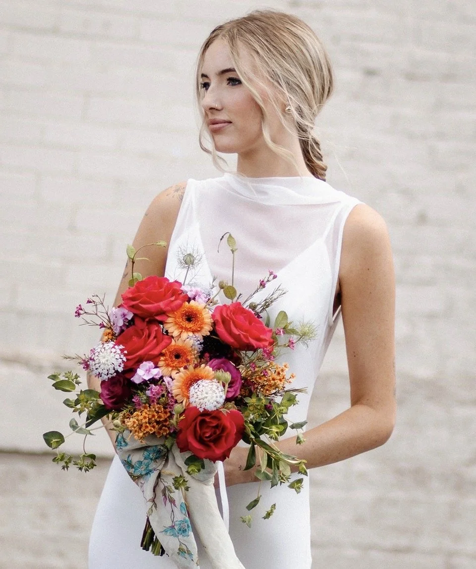 A woman with blonde hair in a loose braid wearing a white dress, holding a colorful bouquet of flowers, standing against a white brick wall.