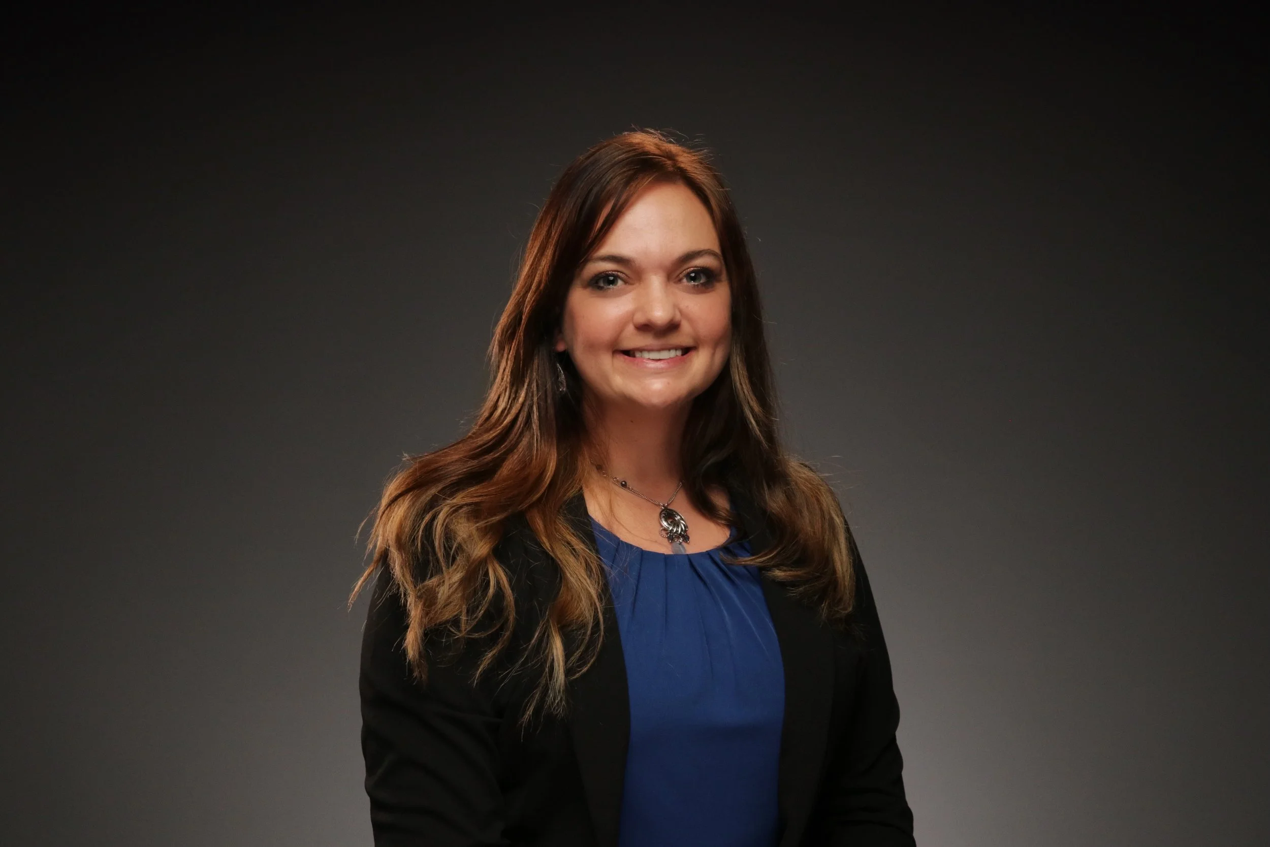 Portrait of a young woman with long wavy brown hair, wearing a black blazer over a royal blue blouse, and a silver pendant necklace, smiling against a dark gray background.