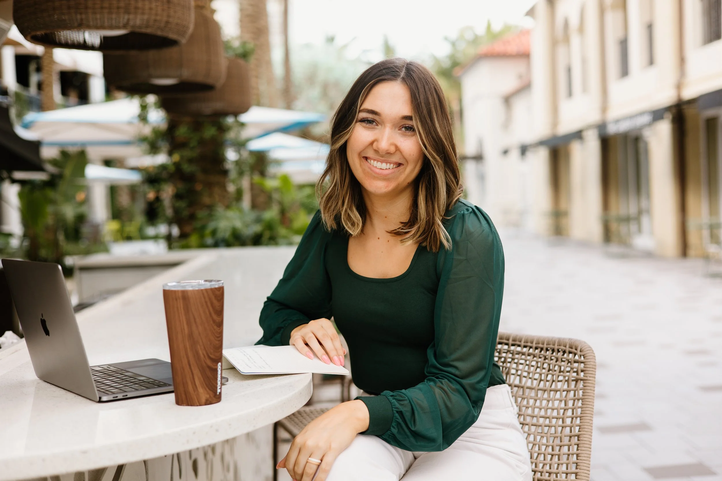 A young woman with shoulder-length brown hair smiling, seated at an outdoor table with a silver laptop and a brown tumbler, wearing a dark green top and white pants, at a cafe with a background of building facades and umbrellas.