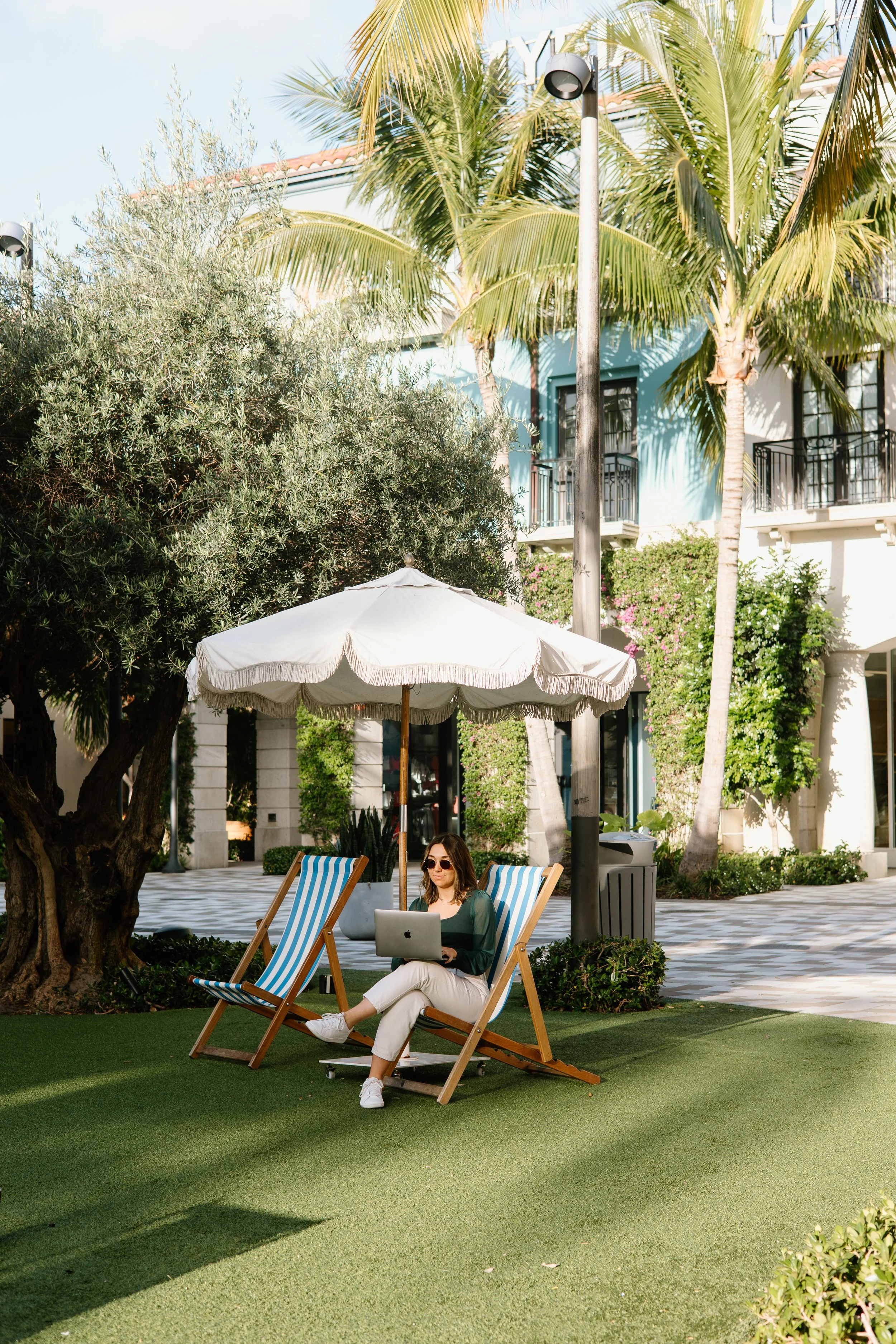 A woman sitting on a striped deck chair outdoors under a white umbrella working on a laptop, with palm trees, bushes, and a building in the background.