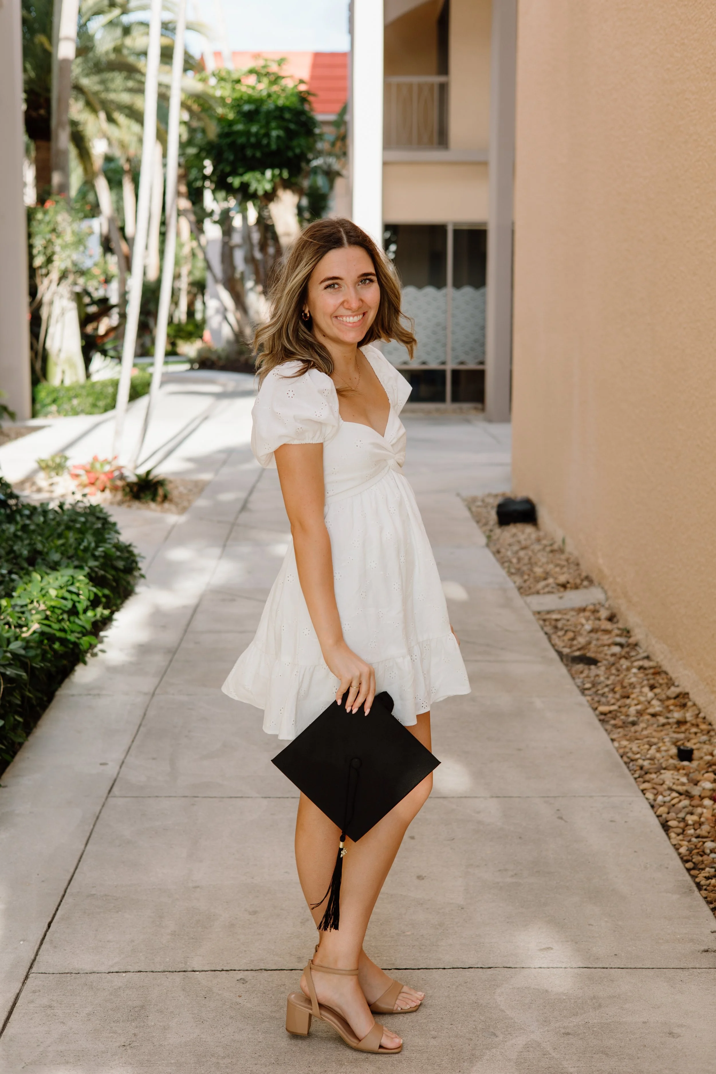 A young woman in a white dress holding a black graduation cap, standing outside on a sidewalk in front of a yellow wall and greenery.