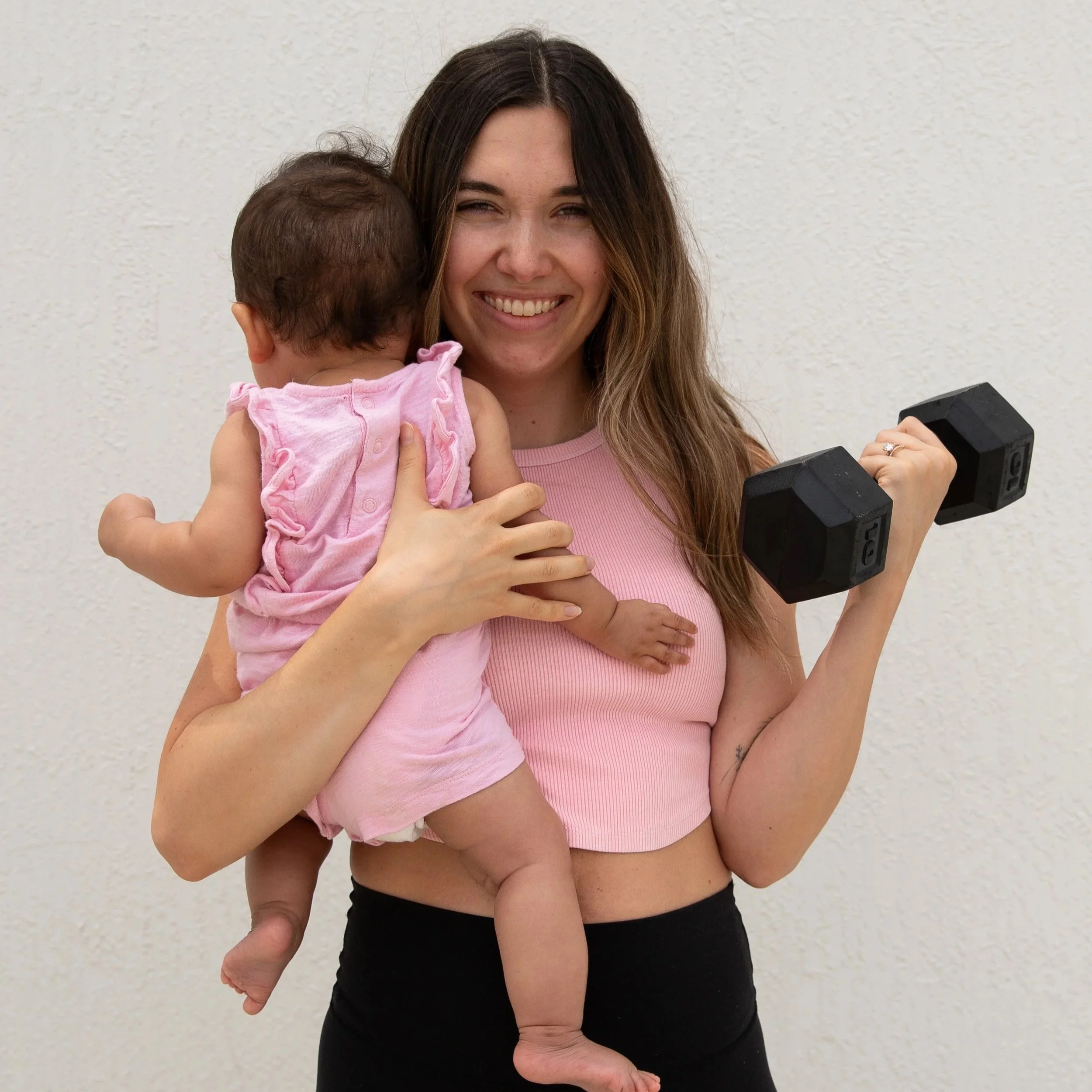 A woman with long brown hair holding a small baby with dark hair and light pink clothing, both against a plain white wall.